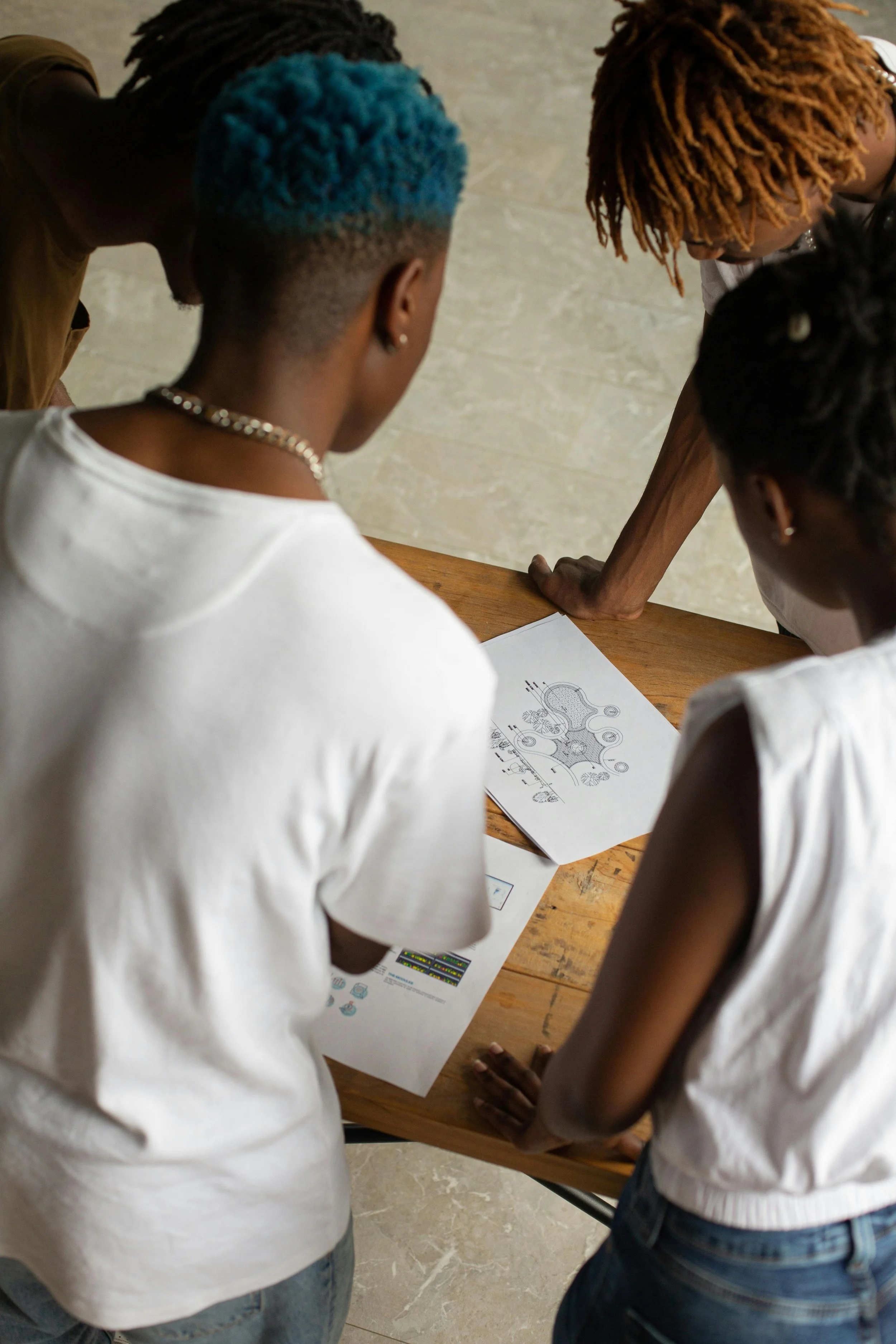Four people gathered around a wooden table, examining printed documents and charts. They are in a casual setting, wearing casual clothing.