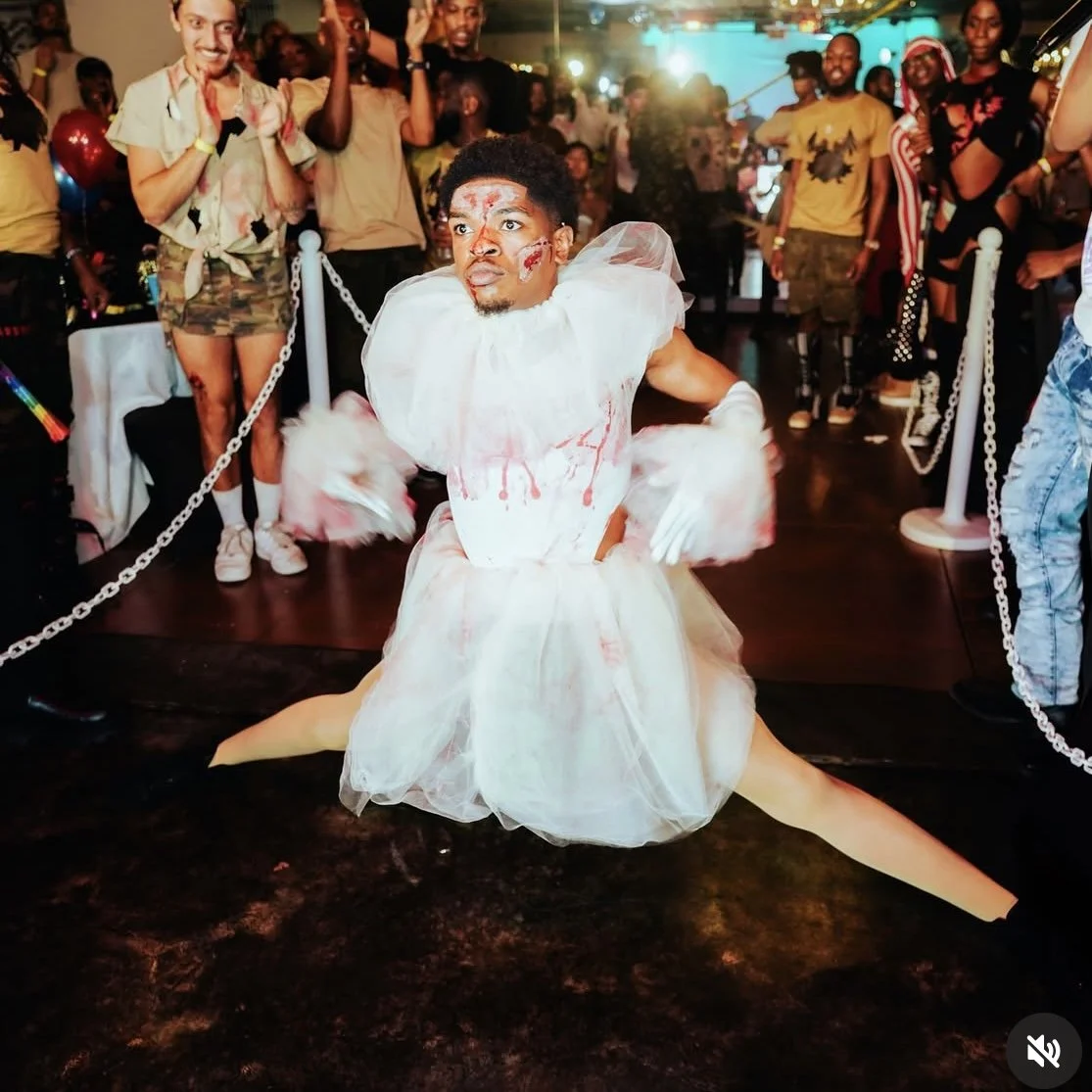 Performer dressed as a bloody bride jumping in a split at a costume party with an audience watching behind a chain barrier.