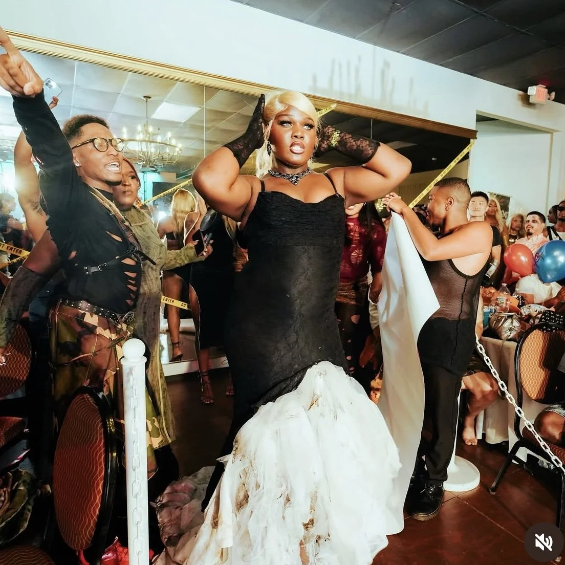 A woman in a black dress and blonde hair poses confidently at a party, surrounded by people, balloons, and festive decorations.