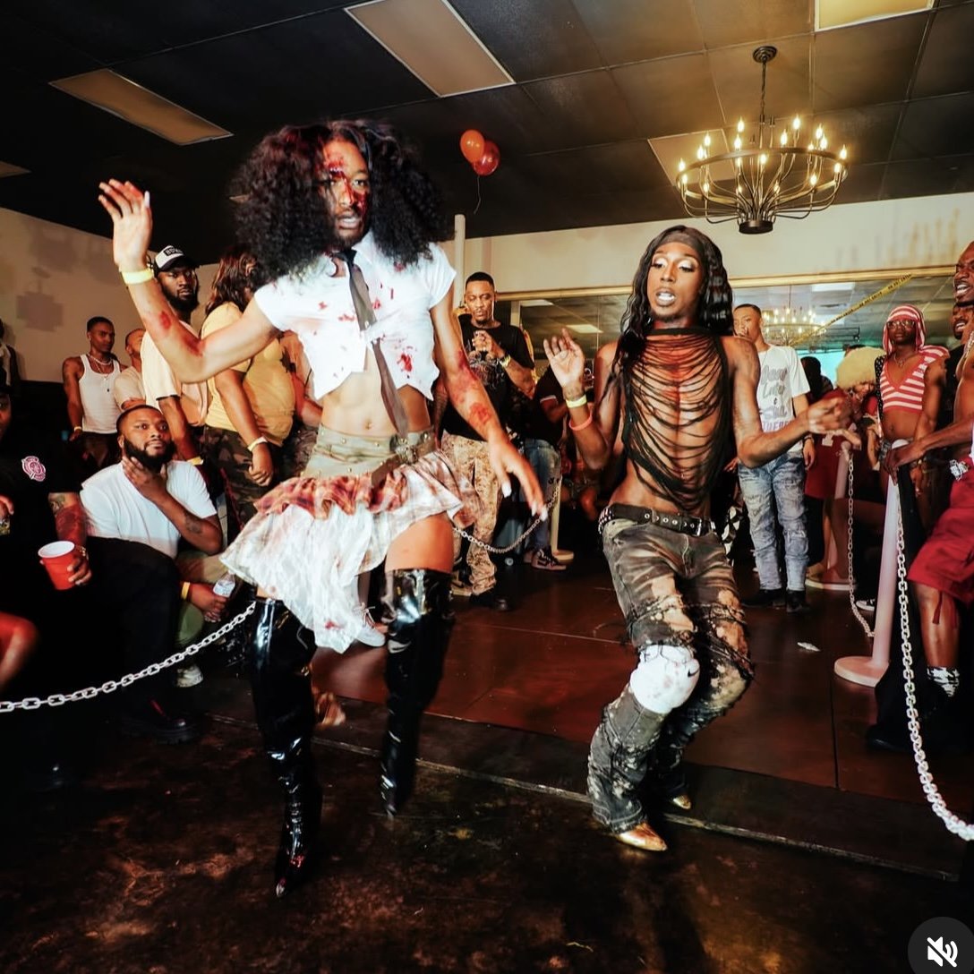 Two women dancing at a party, surrounded by people, with a chandelier and balloons in the background.