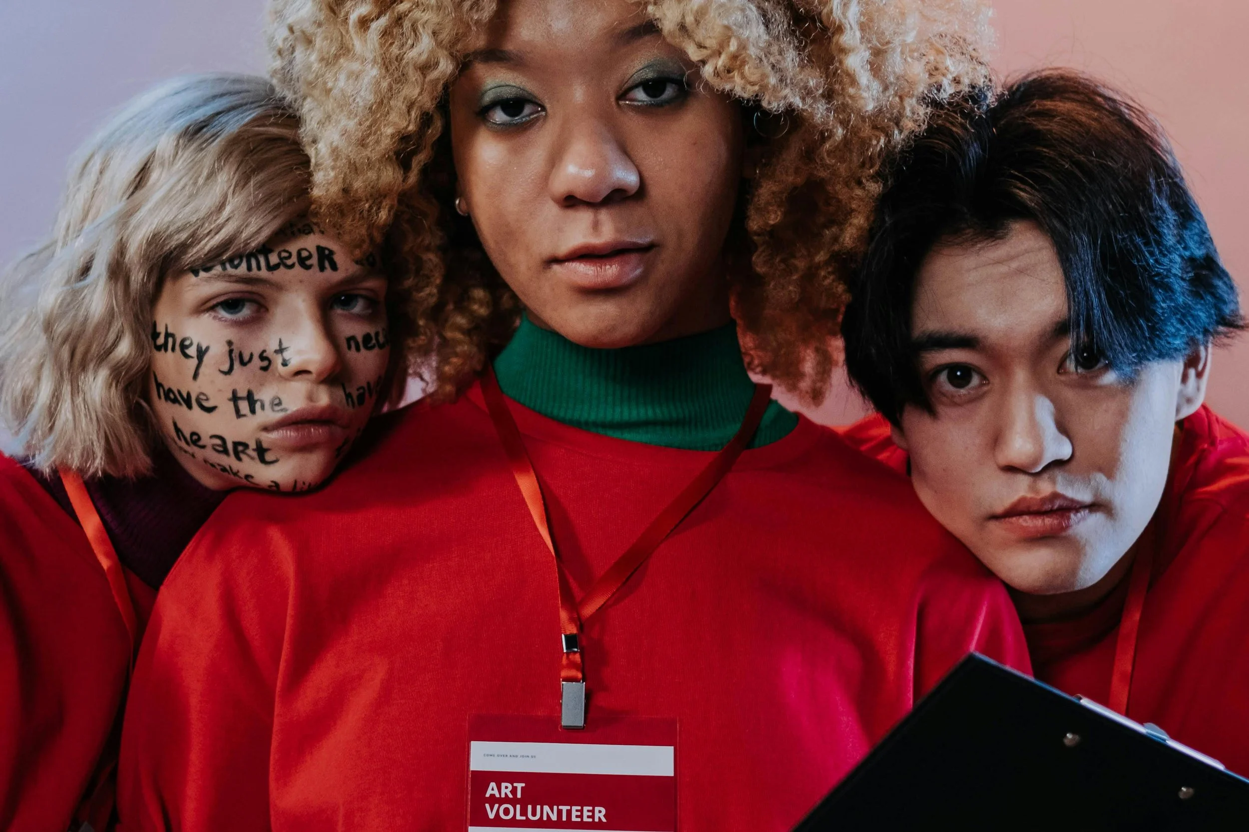 Three volunteers wearing red shirts, with the center woman having curly blonde hair and teal eyeshadow, and the other two individuals flanking her, one with face paint reading 'volunteer' and other with white face paint.