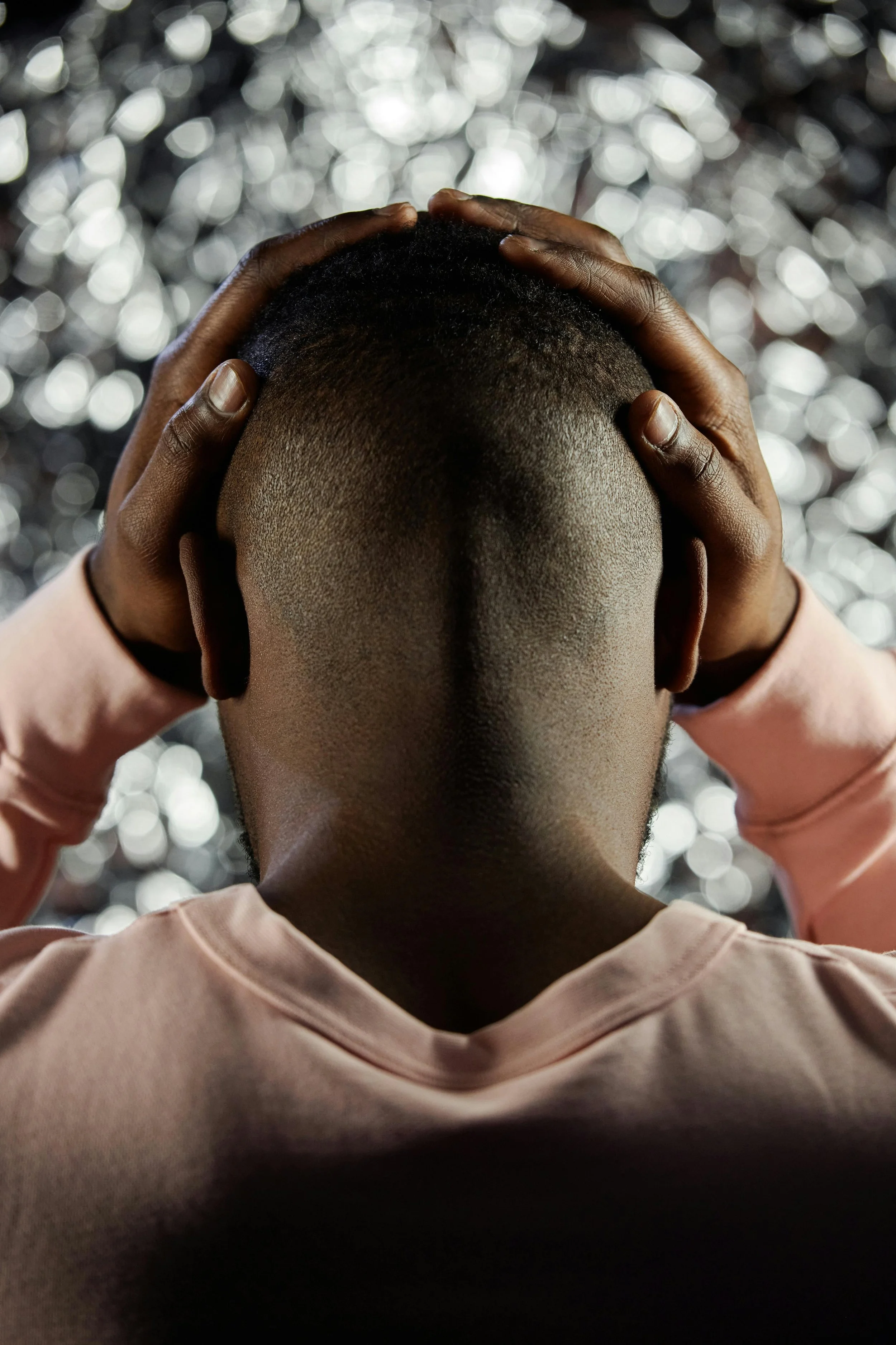 A person with dark skin, holding their head with both hands, cropped at the shoulders, wearing a light-colored top, against a background of blurry bokeh lights.