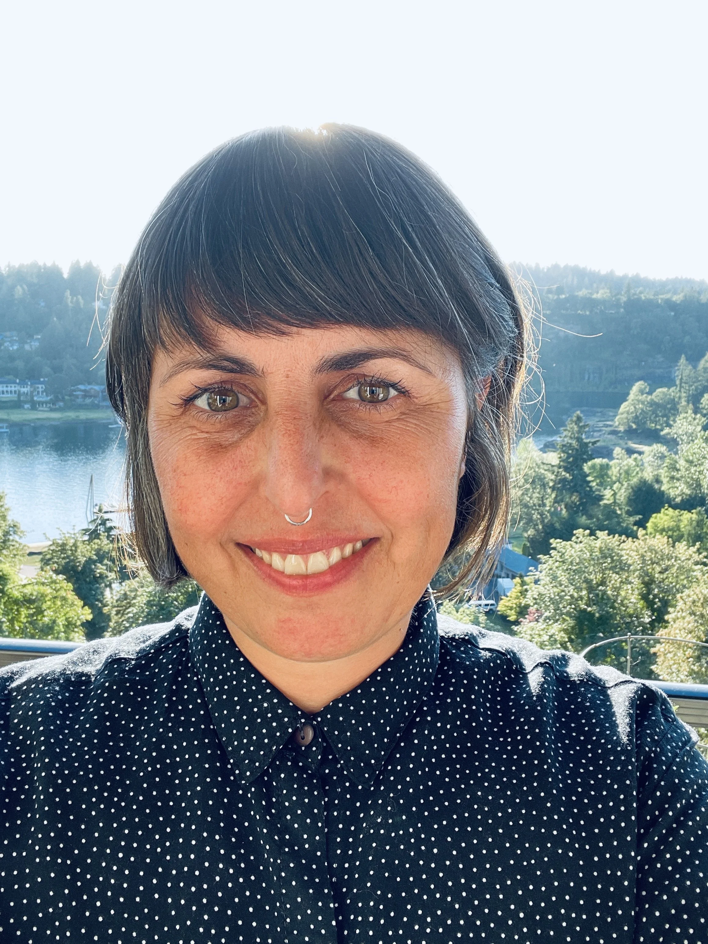 A smiling woman with short dark hair and a nose ring standing outdoors in front of a lake and lush green trees on a sunny day.