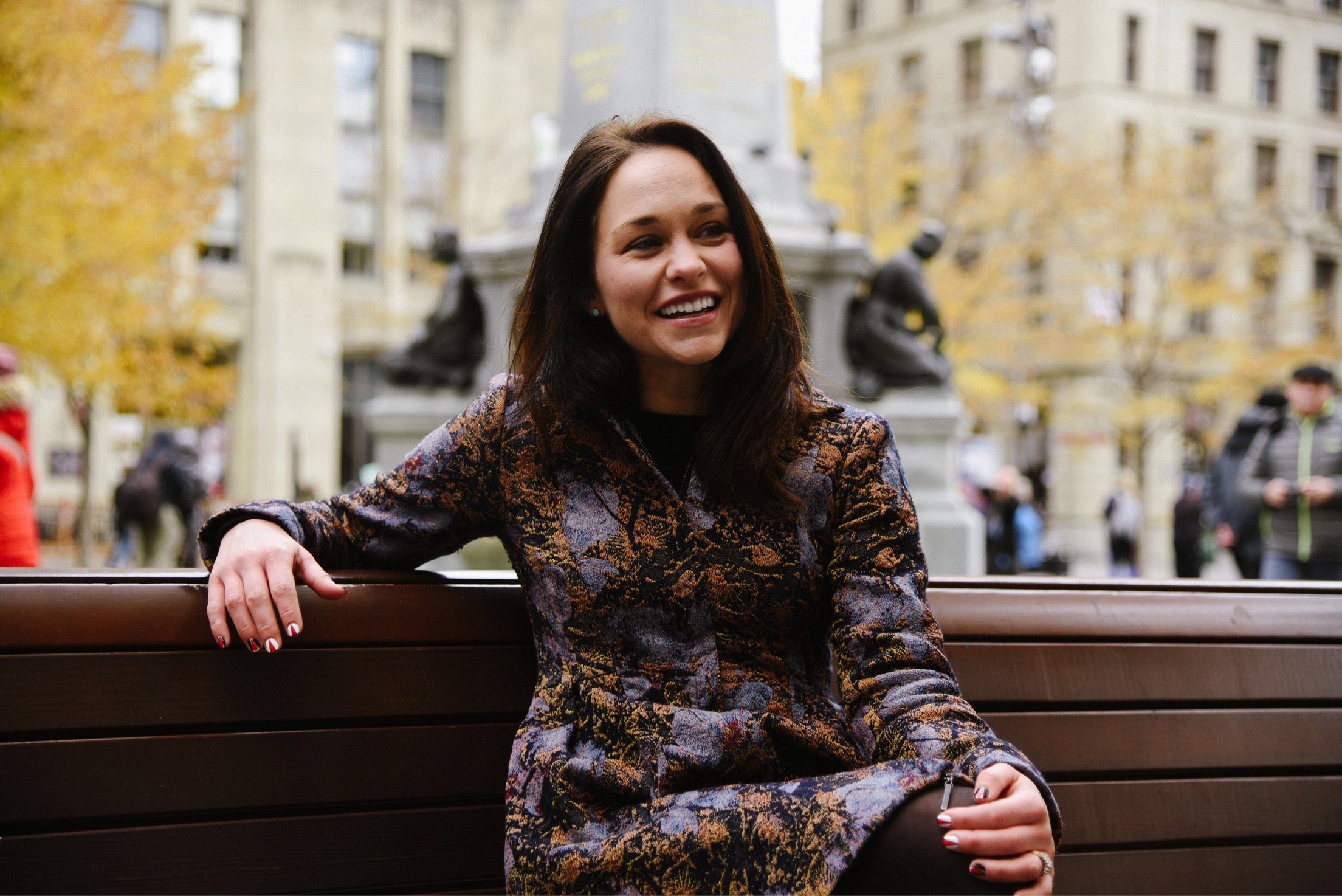 A woman with dark brown hair and a patterned jacket smiling while sitting on a park bench in an urban setting with trees and buildings in the background.