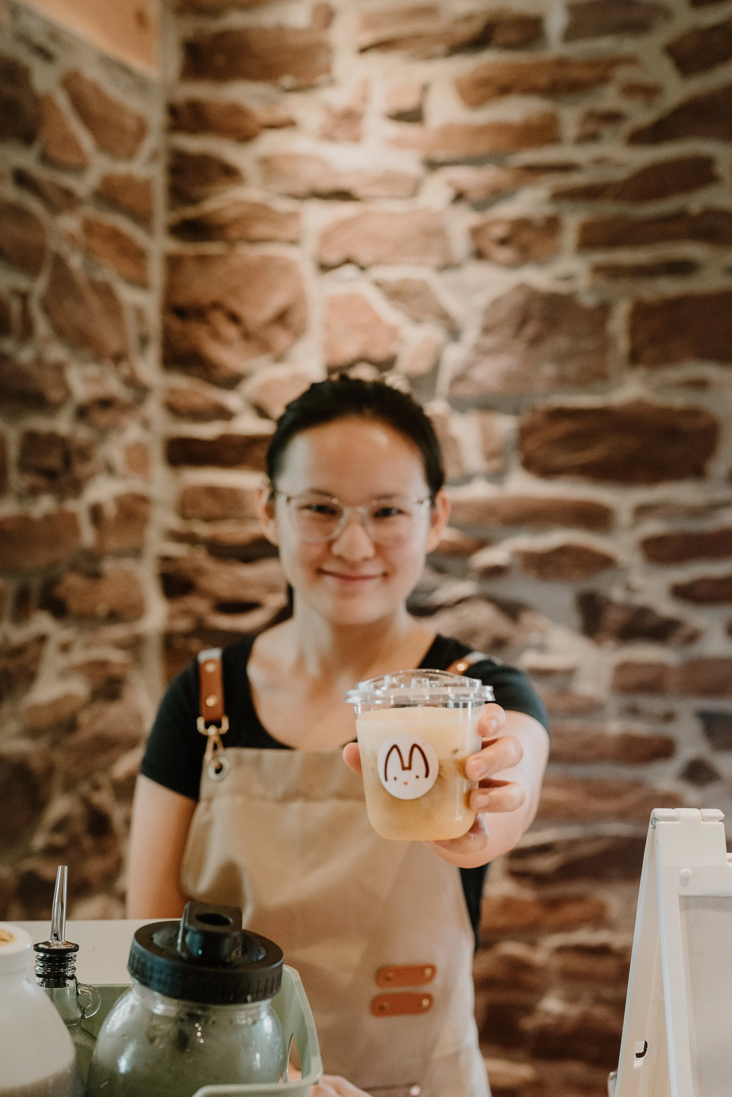 Barista standing behind bar holding out a cold brew drink with branding. Backdrop slightly blurred to show drink in hand.