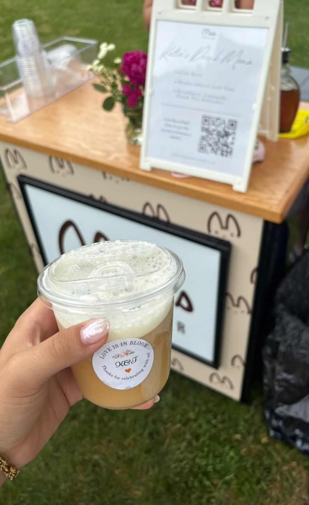 A person's hand holding a mini iced coffee with a custom "Love is in Bloom" sticker, in front of a Minis coffee cart at an outdoor event.