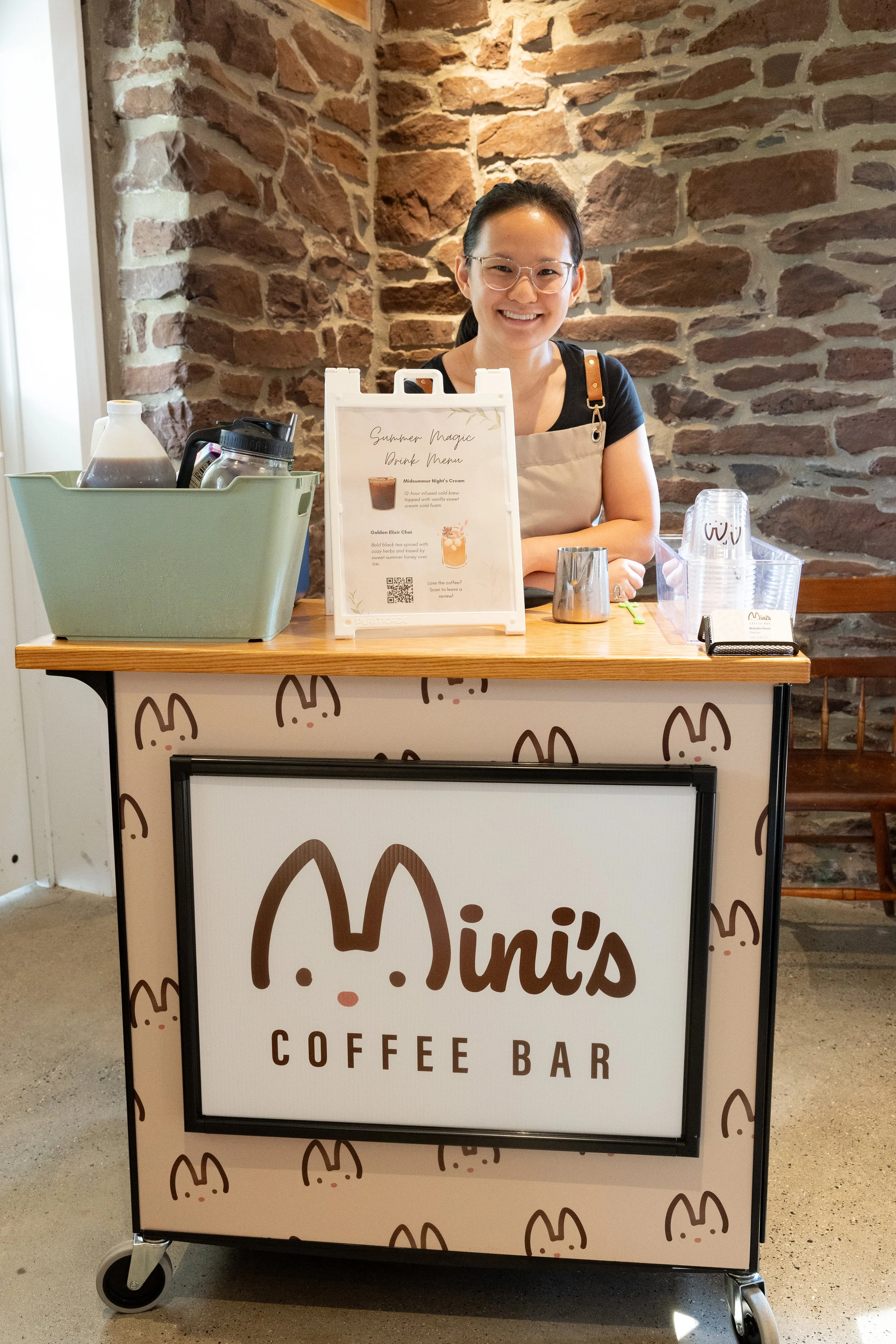Smiling barista standing behind the fully equipped coffee bar with a brick backdrop and warm, overhead lighting. Photo by @luciemarie.photography.