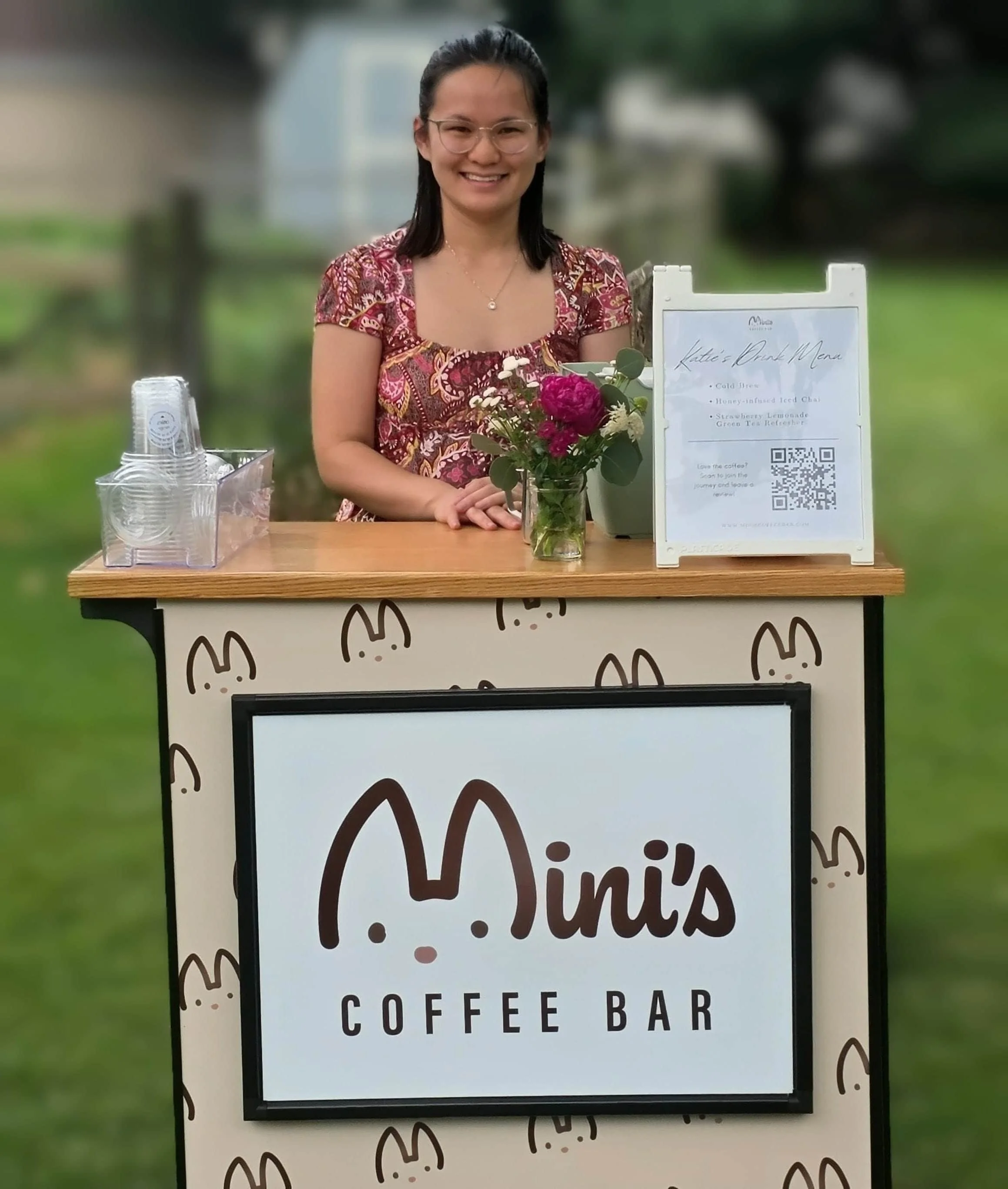 A smiling woman stands behind the Mini's Coffee Bar mobile espresso cart at an outdoor event. The cart features a custom logo sign, a tabletop menu with a QR code, clear plastic cups, and a fresh flower bouquet.