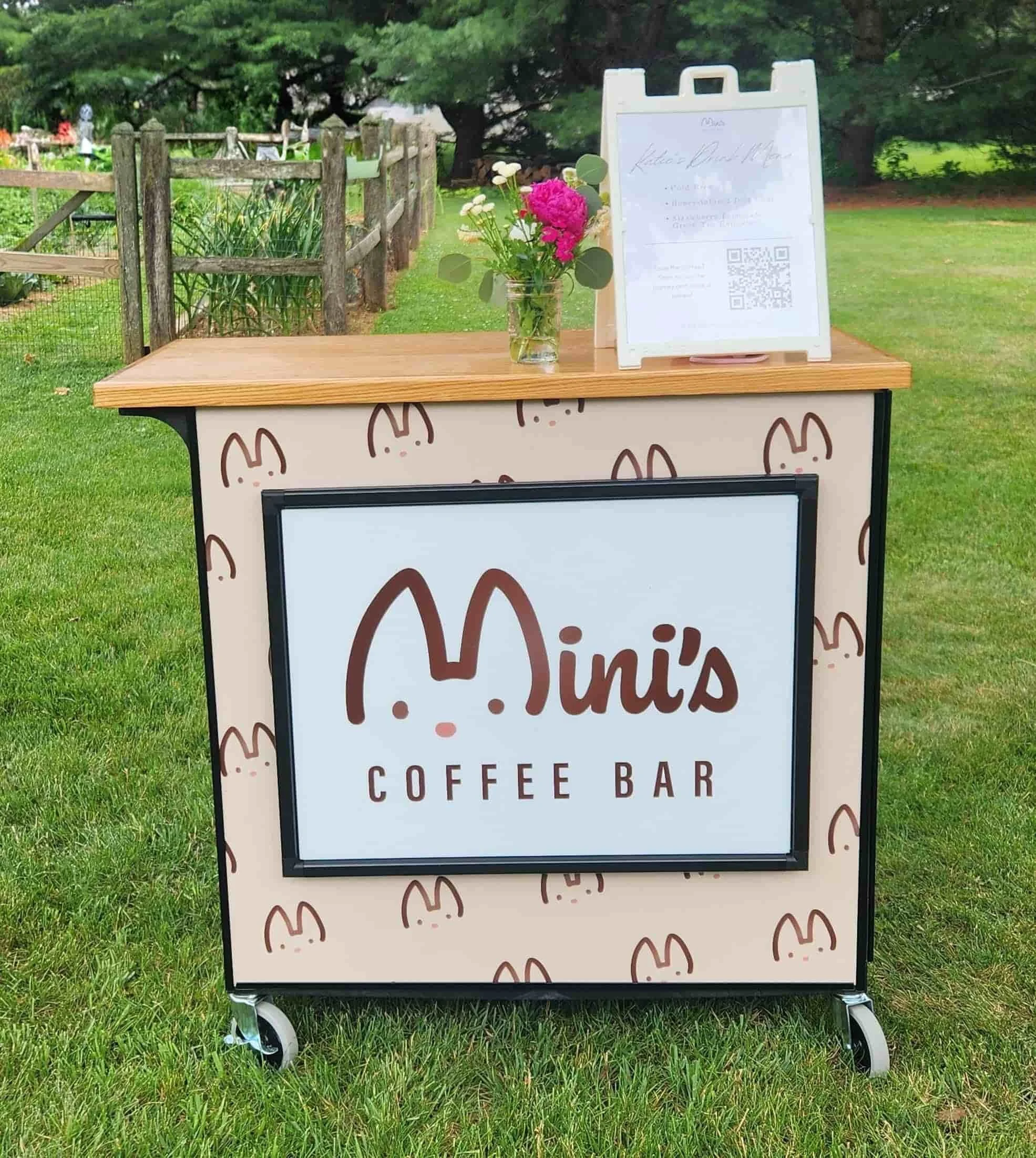 Mobile coffee cart from Mini's Coffee Bar set up outdoors on a lawn, featuring a wooden countertop with fresh flowers, a specialty drink menu sign, and the brand logo on the front.
