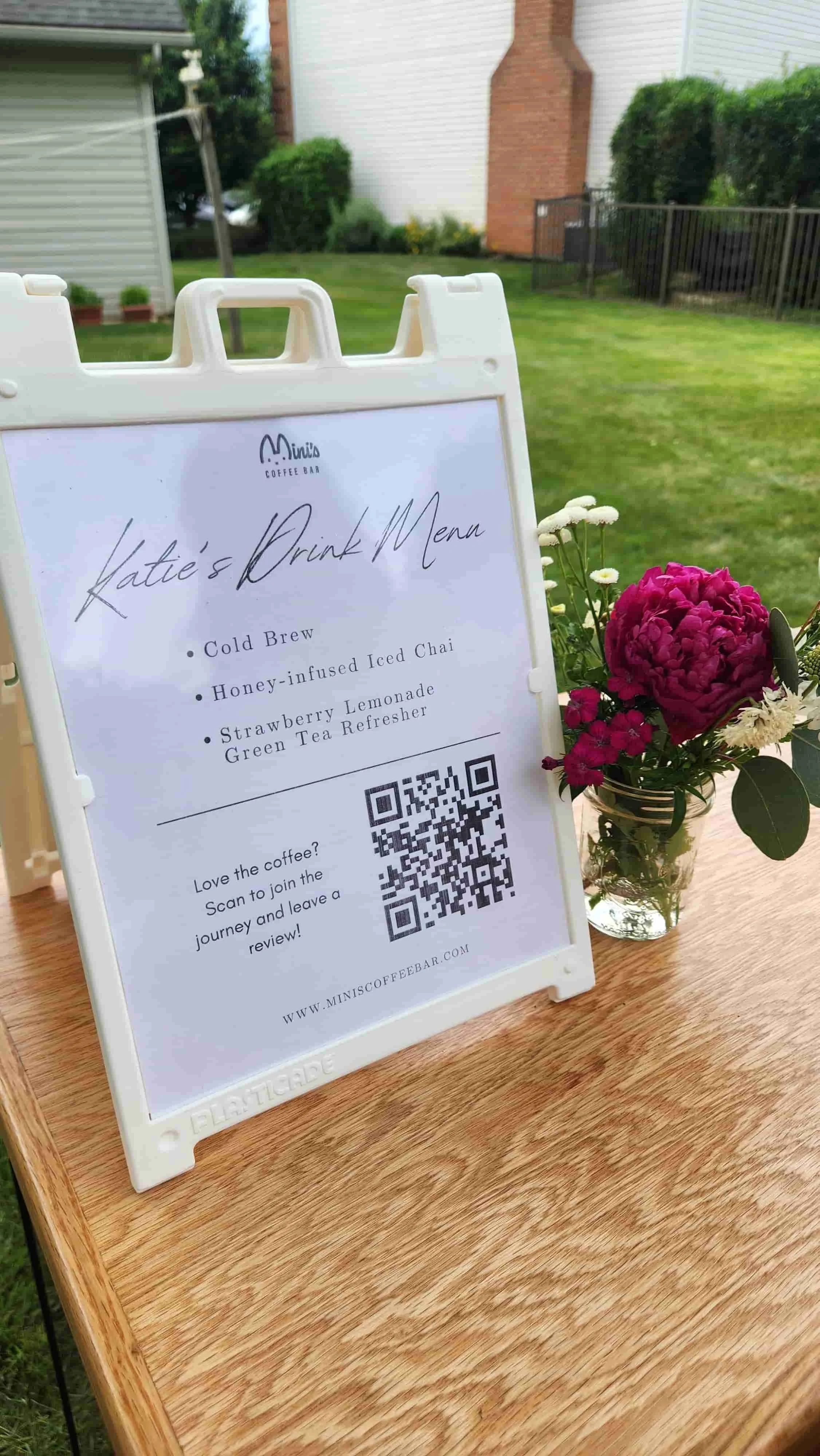 A tabletop menu sign for Mini's Coffee Bar sitting on a wooden counter next to a jar of pink and white flowers. The sign reads "Katie's Drink Menu" and features a QR code inviting guests to leave a review.