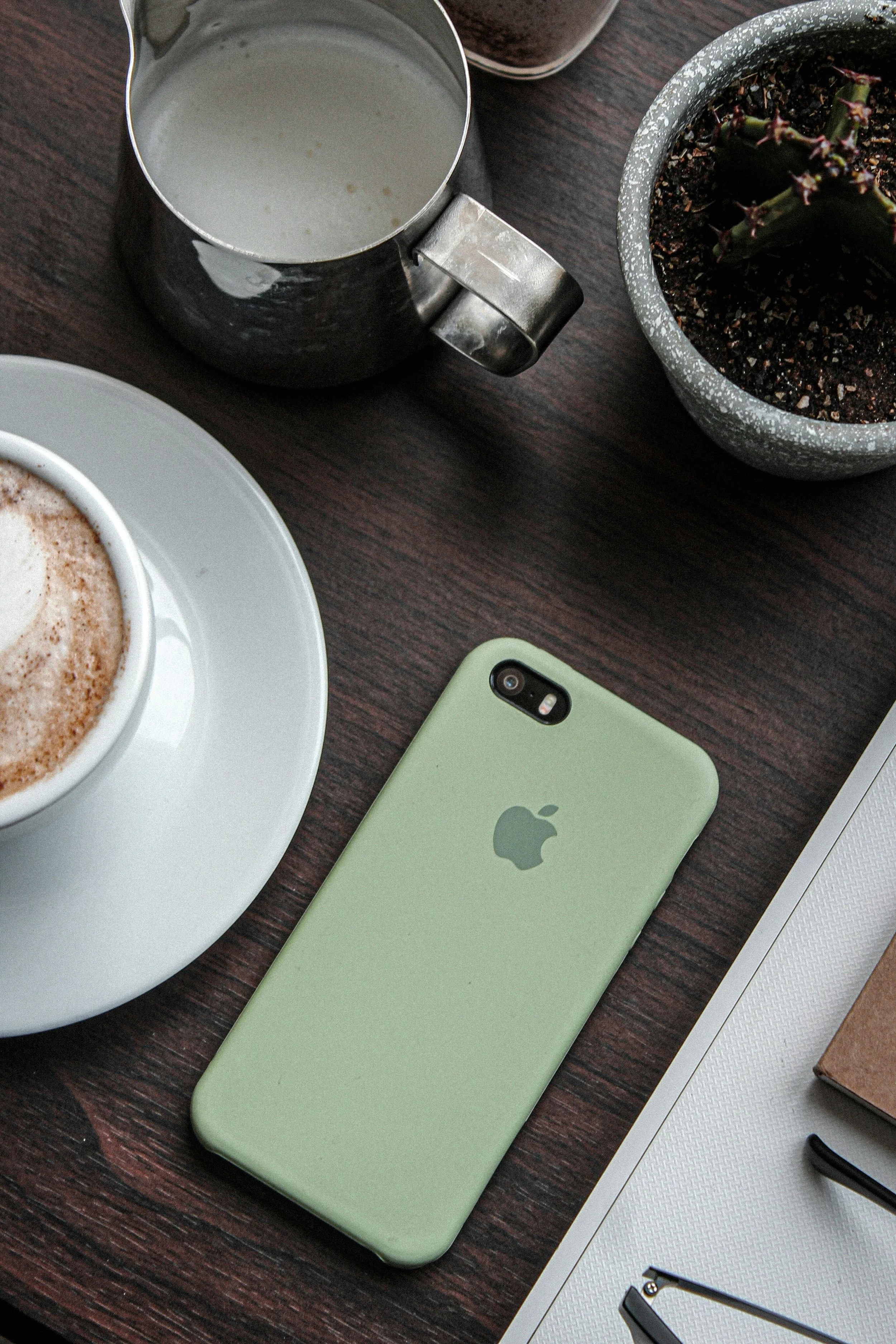 Top-down view of a dark wooden desk featuring a white coffee cup with a foam design, a green iPhone, a silver coffee pot, a gray planters with a cactus, a part of a laptop keyboard, and a small orange box.