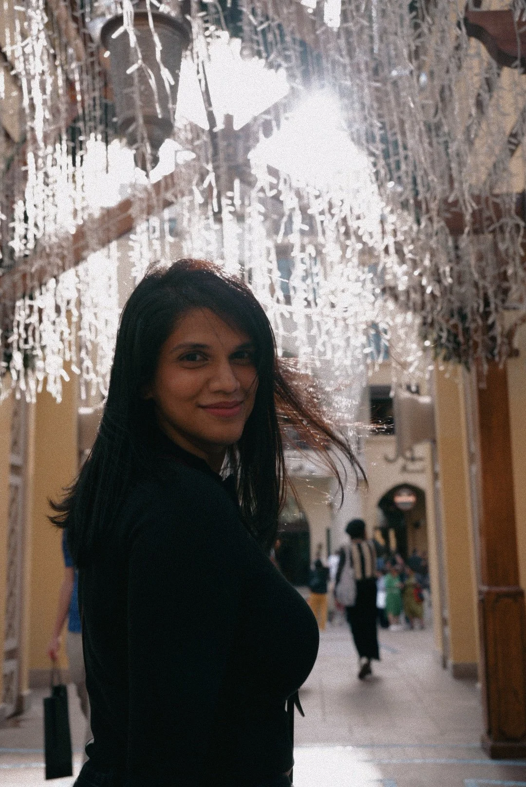 A young woman with dark hair smiling at the camera inside a shopping mall decorated with white hanging ornaments and bright lights.
