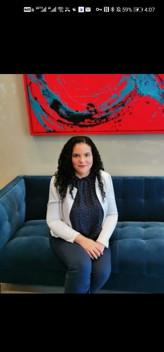 A woman with curly dark hair sitting on a blue velvet sofa, wearing a white blazer over a black polka dot blouse, with a colorful red and blue abstract painting behind her.
