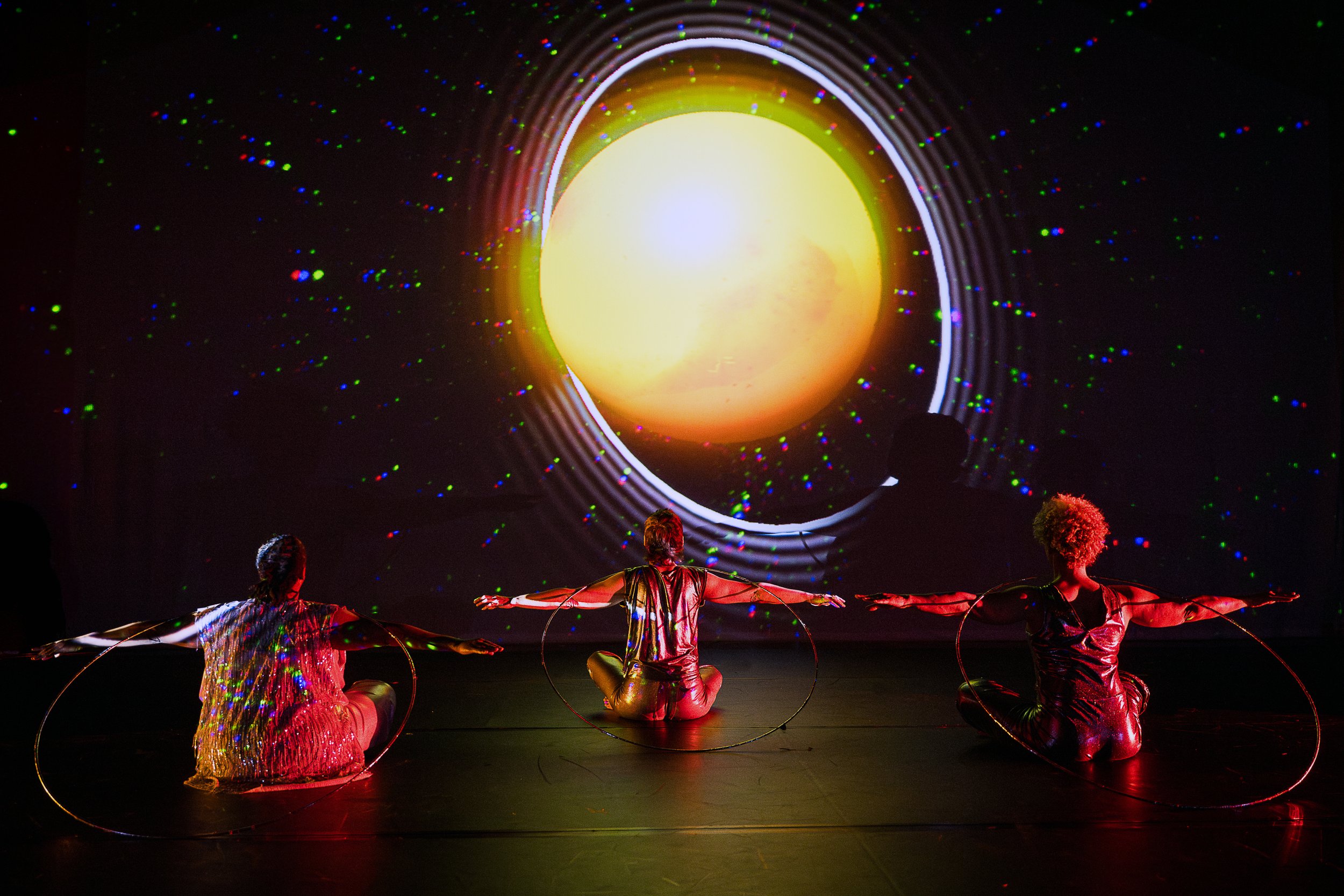 Kayla Hamilton, Tatiana Cholewa, and Alice Sheppard sit cross-legged on the floor, their backs to the camera. They extend their arms wide, energy radiating from their fingertips. They balance a thin silver hoop on their neck. Photo Cherylynn Tsushima