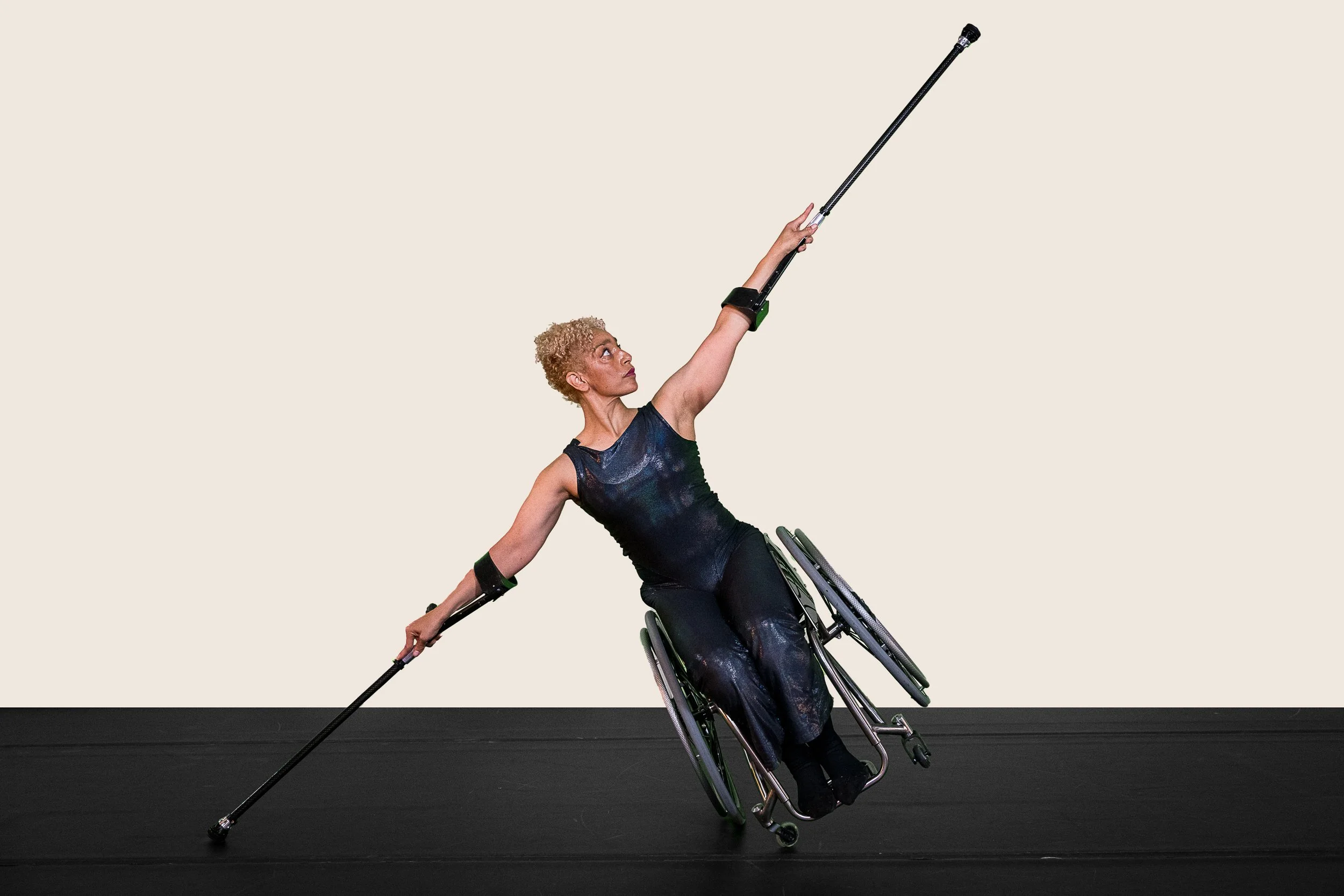Alice Sheppard lifts and tucks her knees to her chest as she balances on her crutches. Her wheelchair rests on the floor behind her. Alice is a multiracial Black femme with coffee-colored skin and blonde curly hair piled atop her head.