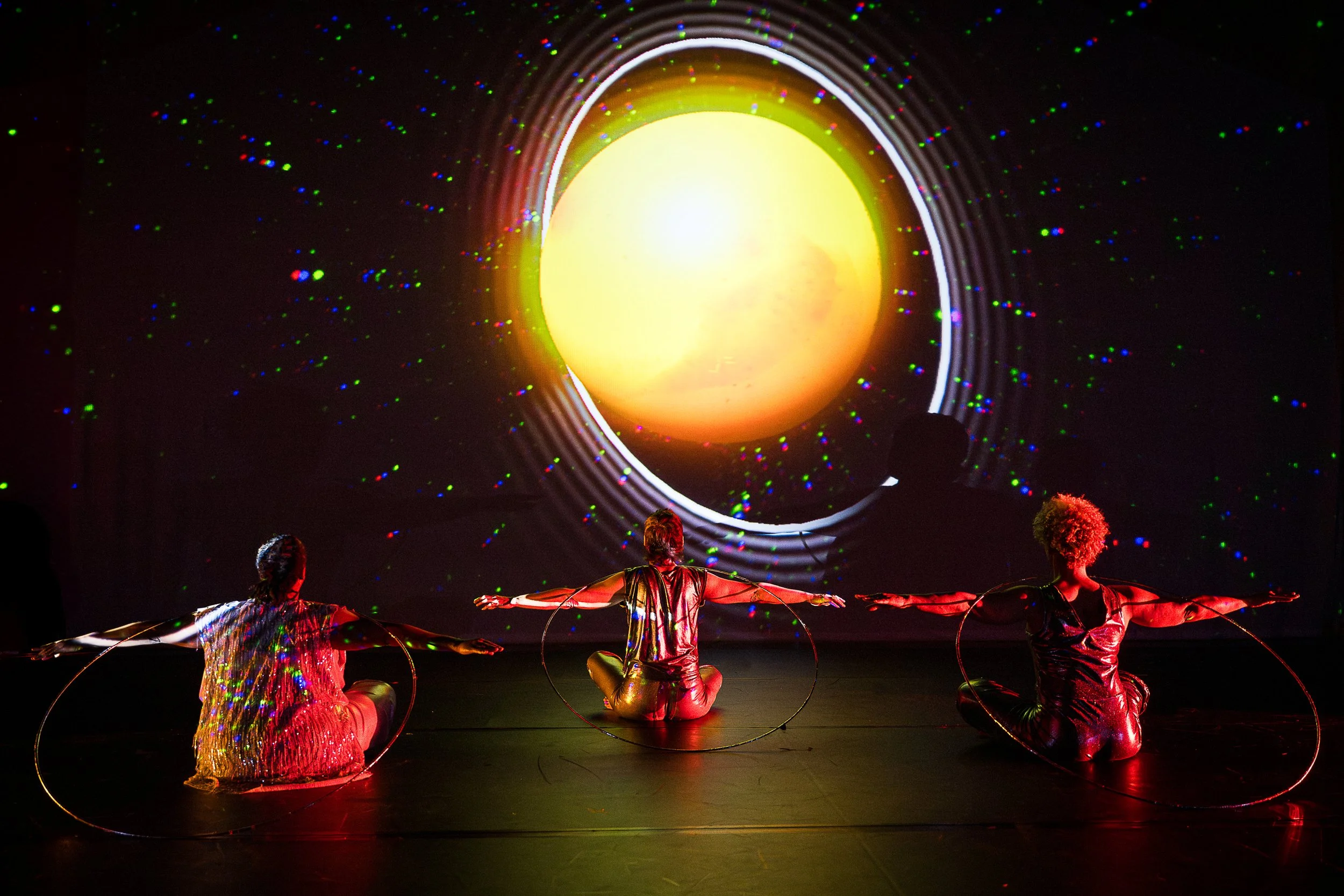 three performers sitting on stage, arms outstretched, facing a multicolor backdrop