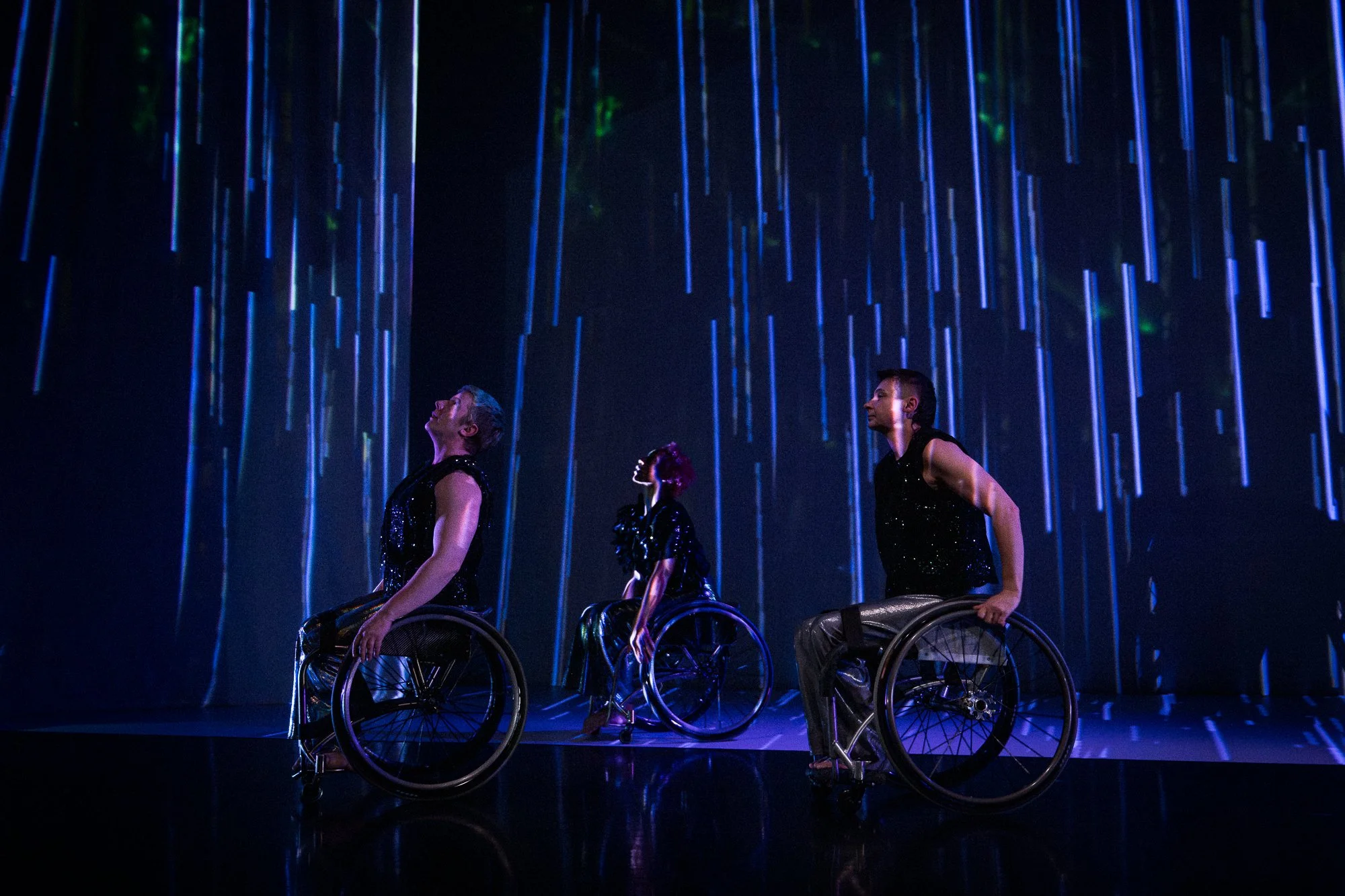 three performers in wheelchairs turned left in front of a blue backdrop
