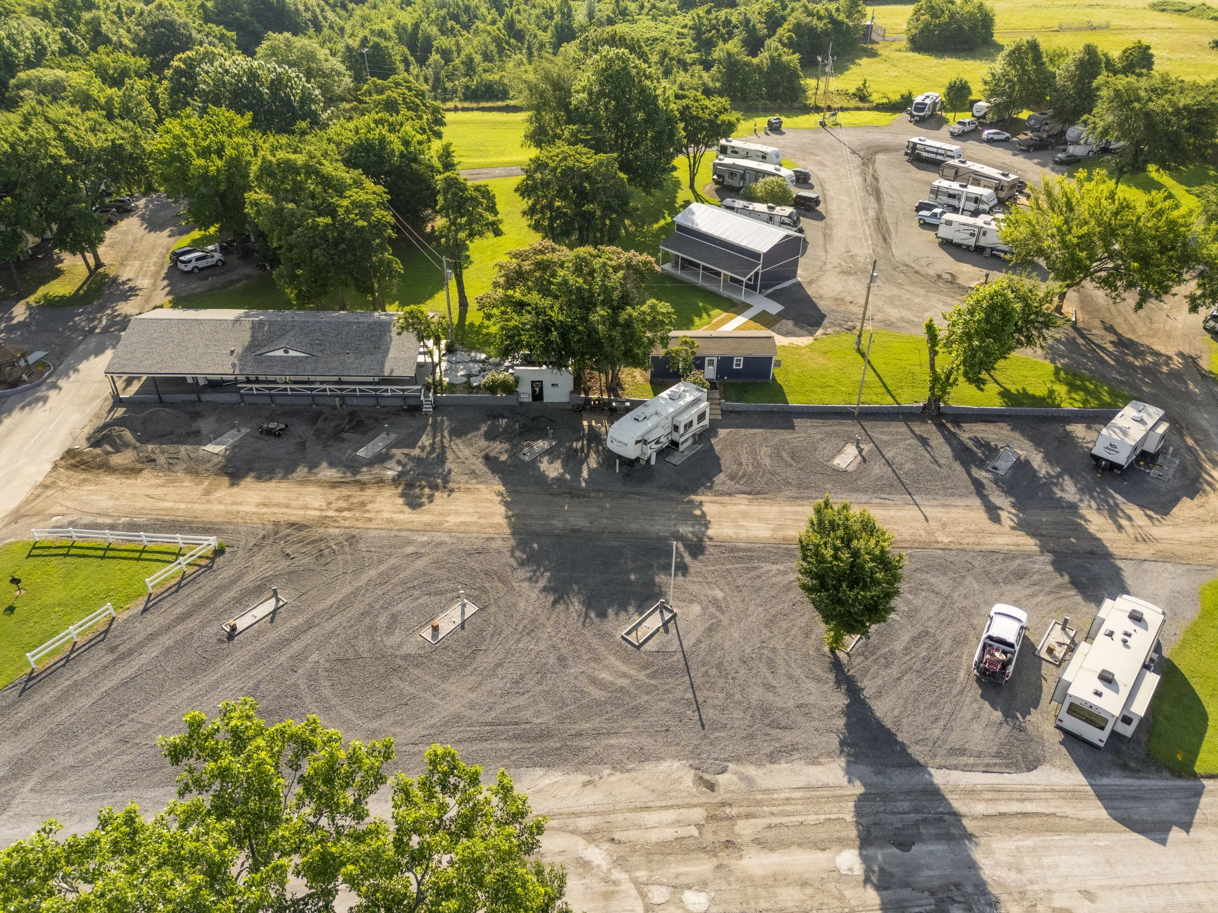 An aerial view of a campground with multiple RVs and trailers parked among green trees and grass. There are some small buildings, a gravel parking lot, and a construction area with dirt and unpaved roads.