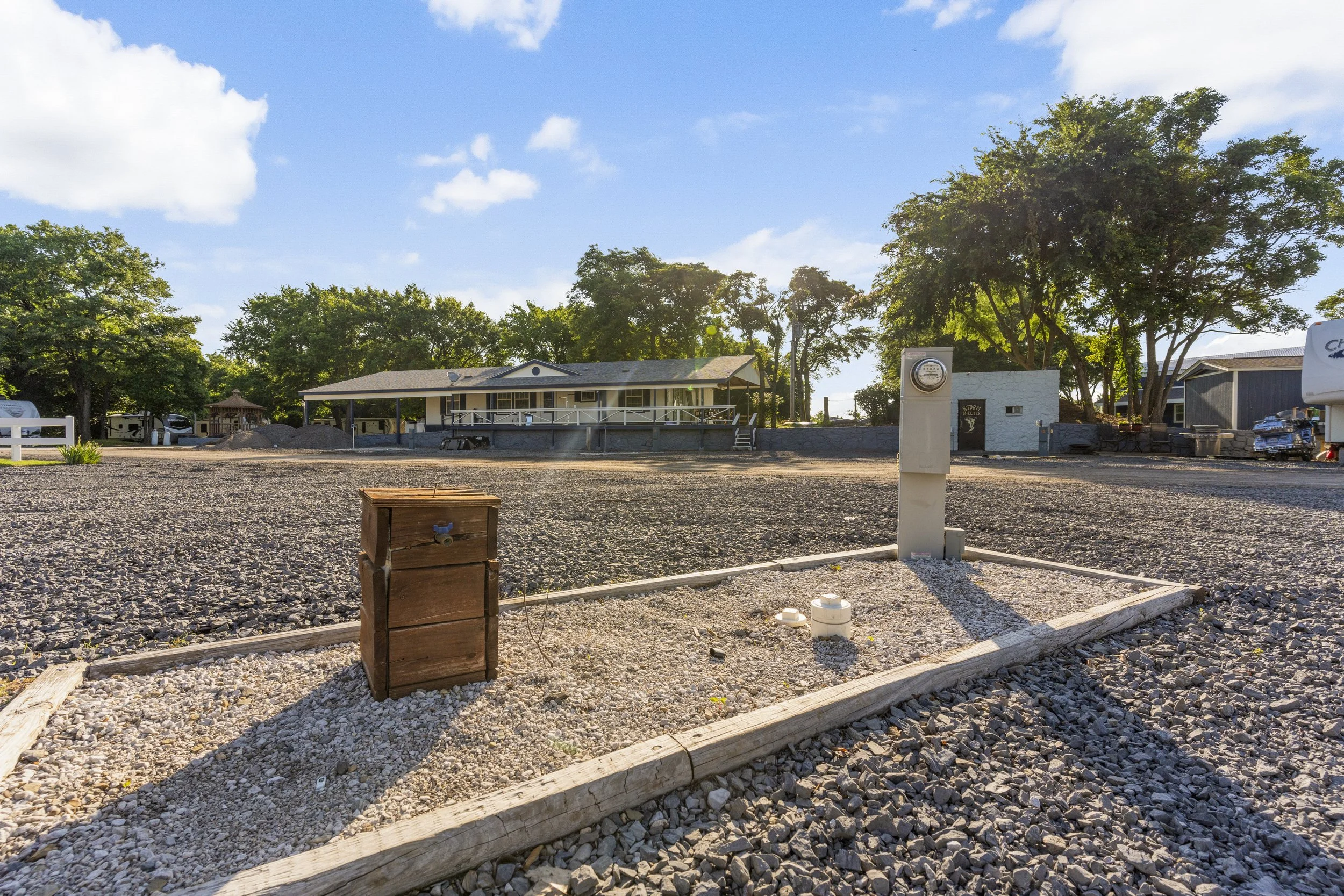A gravel lot with a small wooden box in the foreground and a white utility box in the middle ground. In the background, there is a building with a porch, surrounded by trees, and a partly cloudy sky.