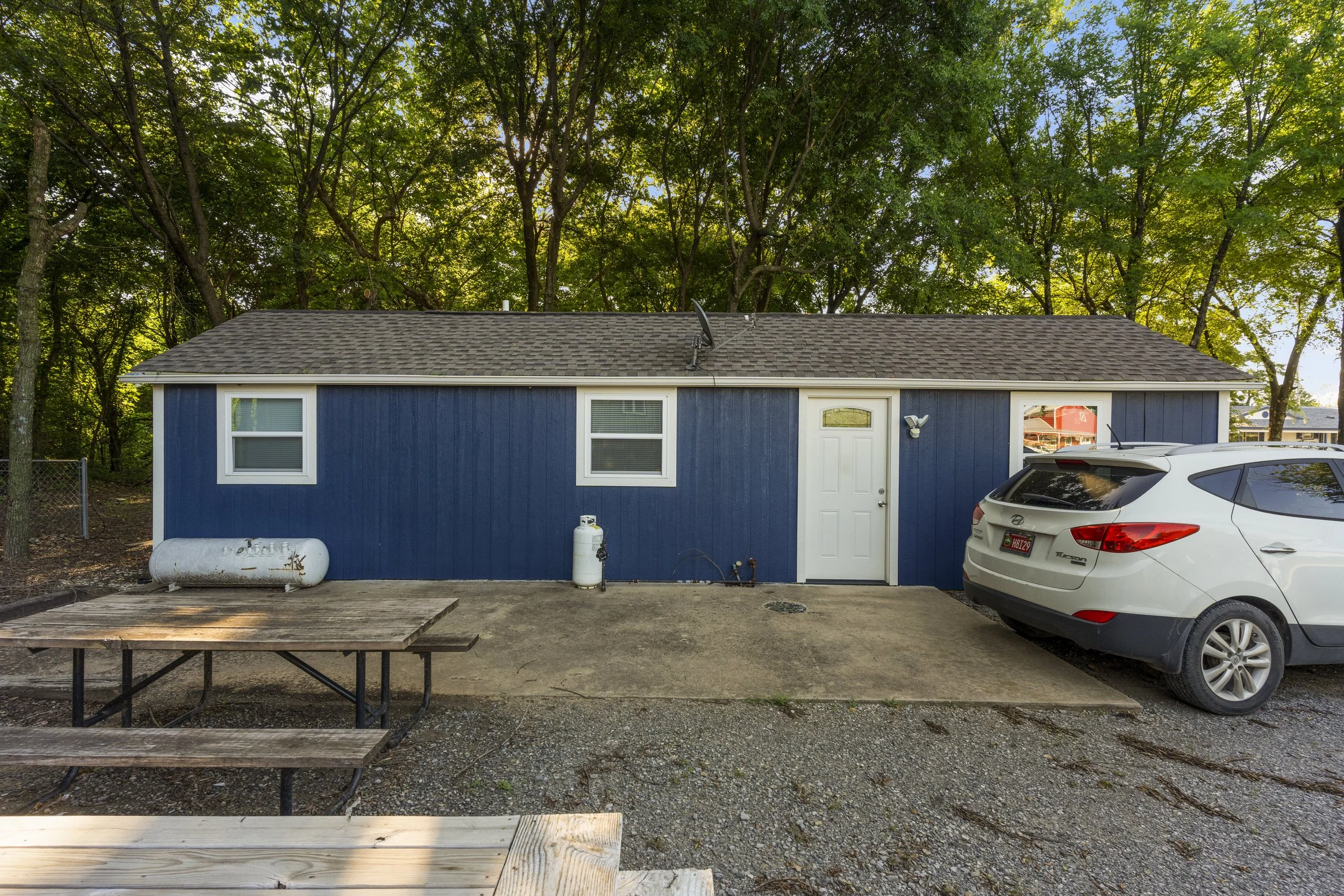 A small blue house with white trim features three windows, a white door, and a satellite dish on the roof. In front of the house, there is a wooden picnic table and a white SUV parked nearby. The scene is set in a wooded area with tall green trees in