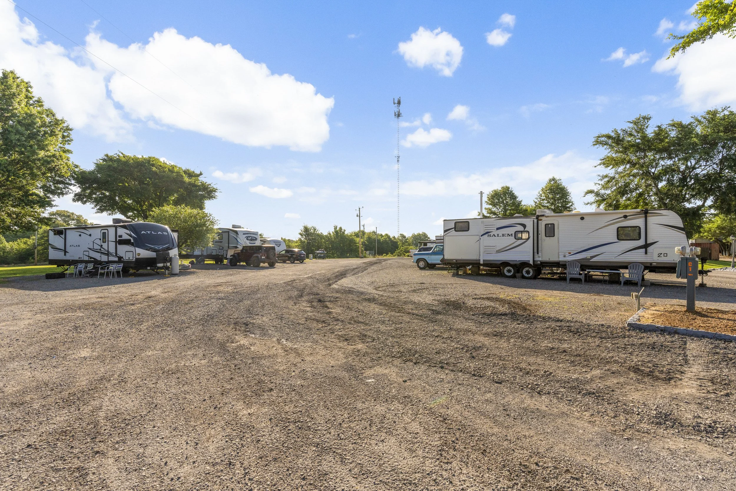 A dirt parking lot with several RVs and vehicles parked under a clear blue sky with a few white clouds, surrounded by green trees.