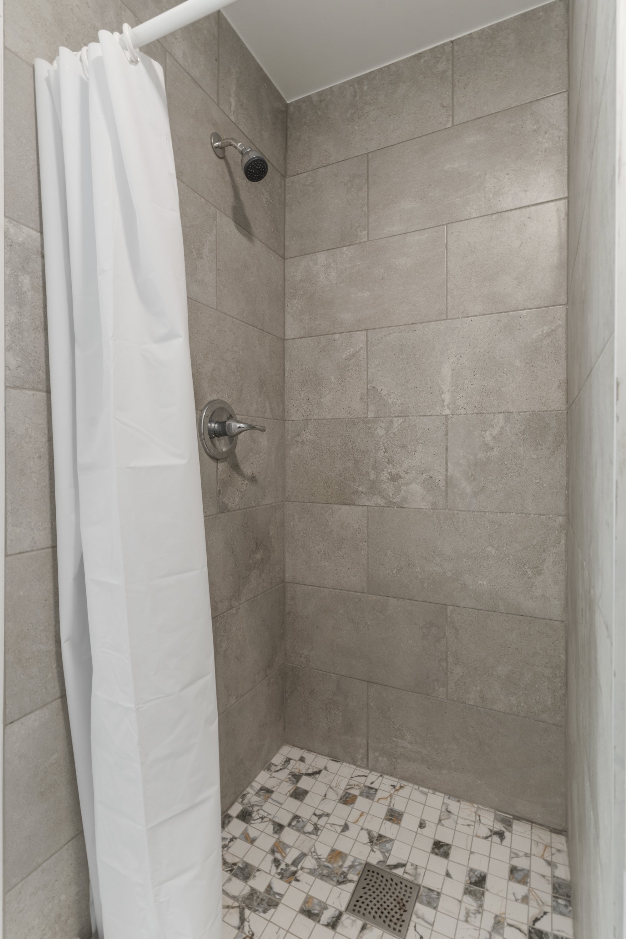 A walk-in shower with beige tiled walls, a white shower curtain, a silver showerhead, a silver water control knob, and a floor with white and gray marble tiles and a metal drain.