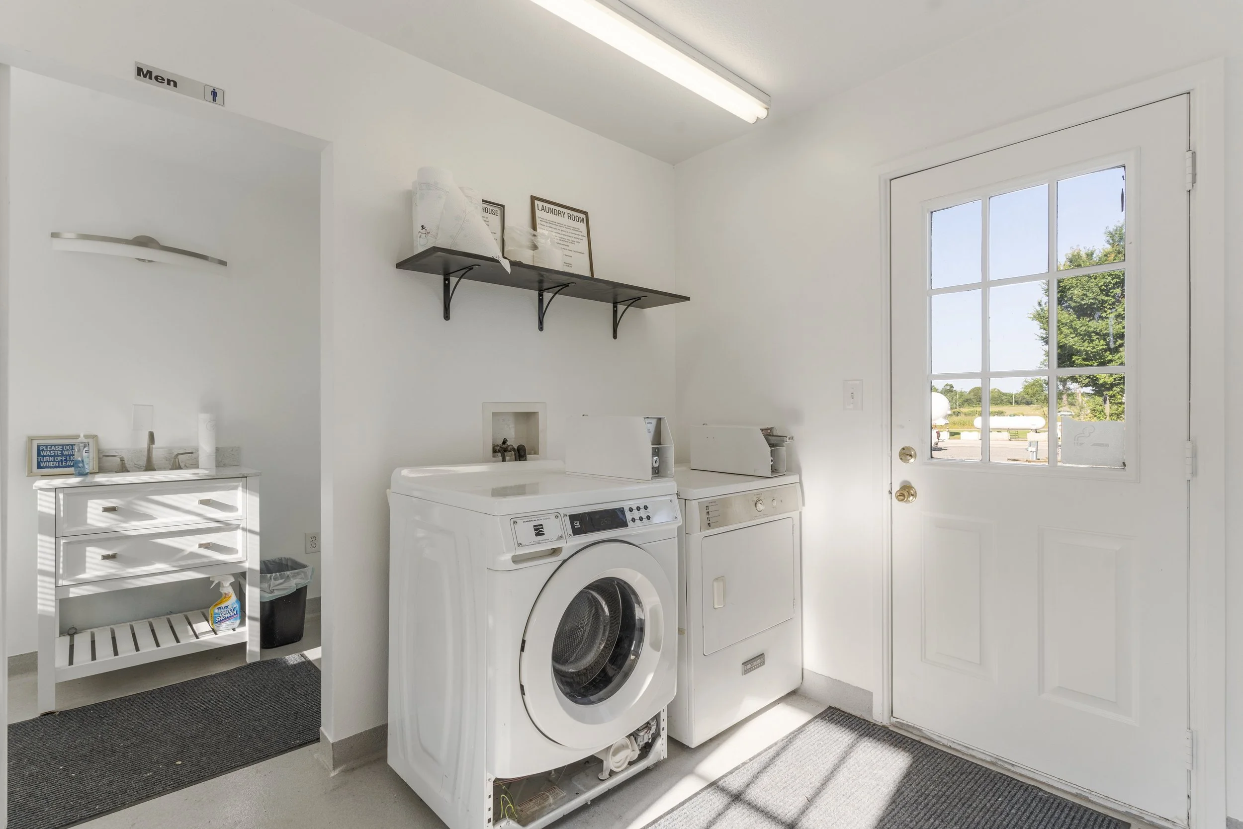 Laundry room with a washing machine, dryer, white shelving, and a door with a window showing outdoor scenery.