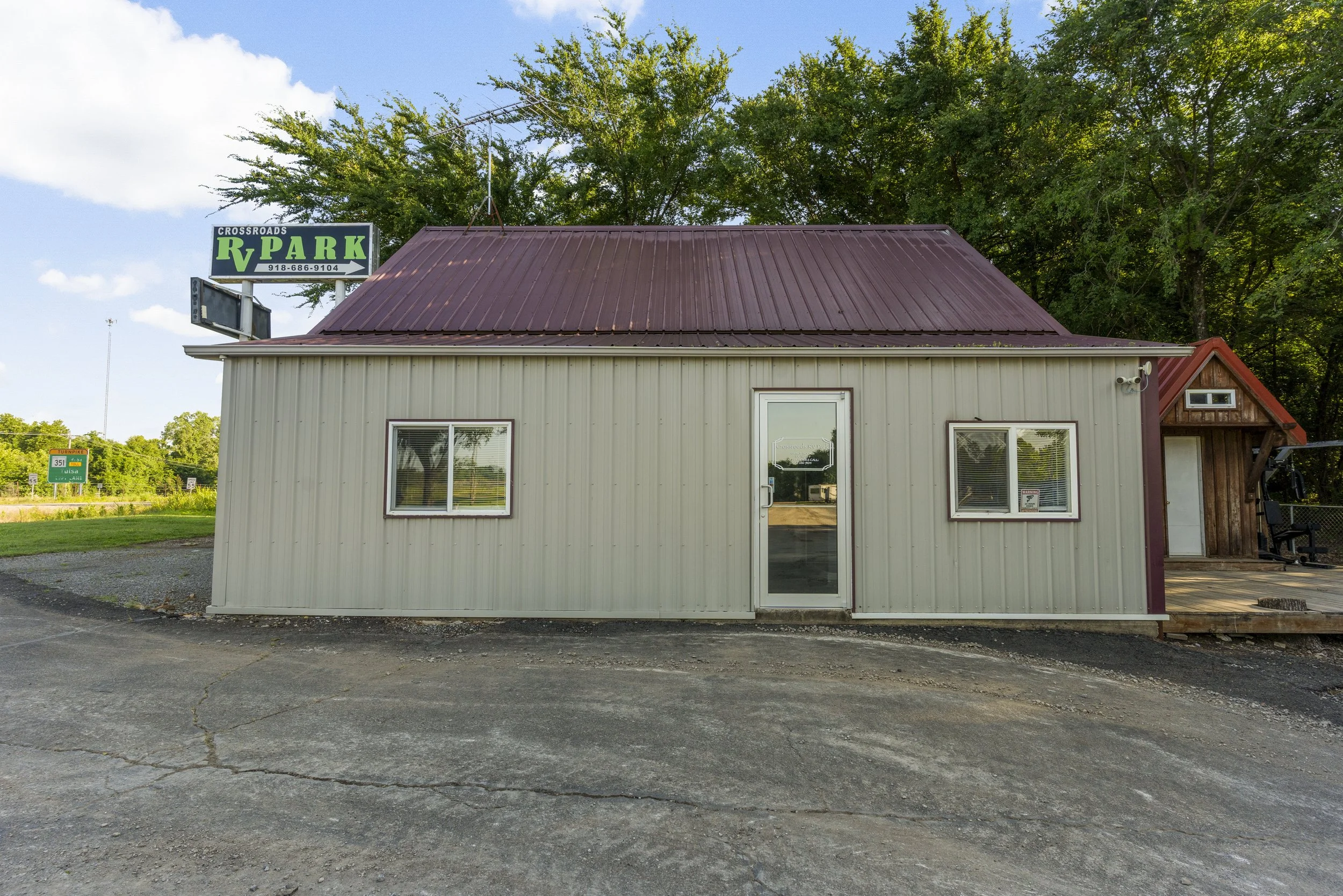 A small beige building with a red metal roof and white door, surrounded by green trees, with a sign for Crossroads RV Park visible above the roof.