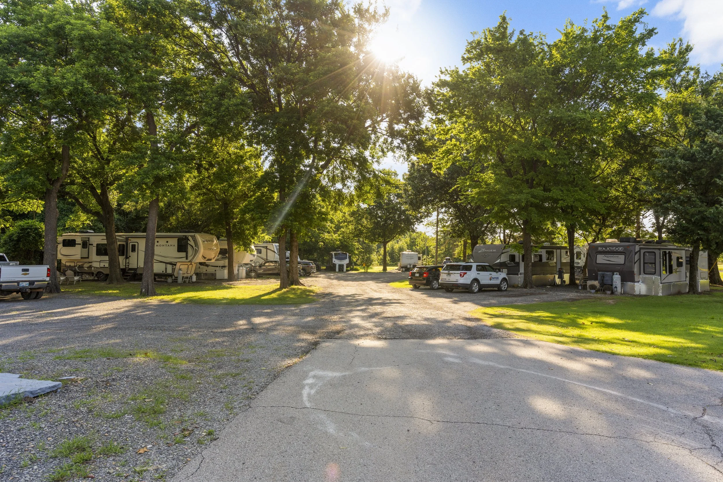 Sun shining through green trees at an RV campground with several parked RVs, trucks, and cars on a gravel lot.