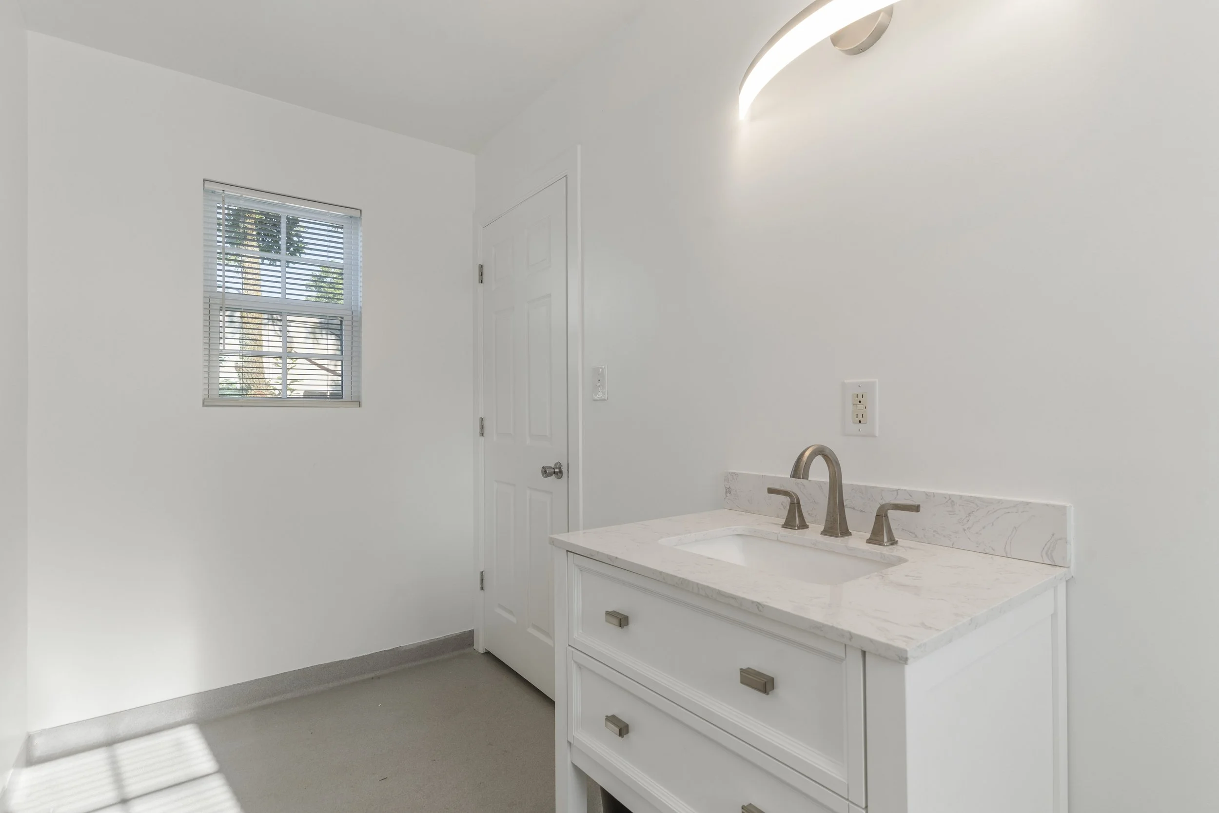 A white bathroom vanity with drawers and a marble countertop, stainless steel faucet, a window with blinds, and a door, in a brightly lit room with white walls.