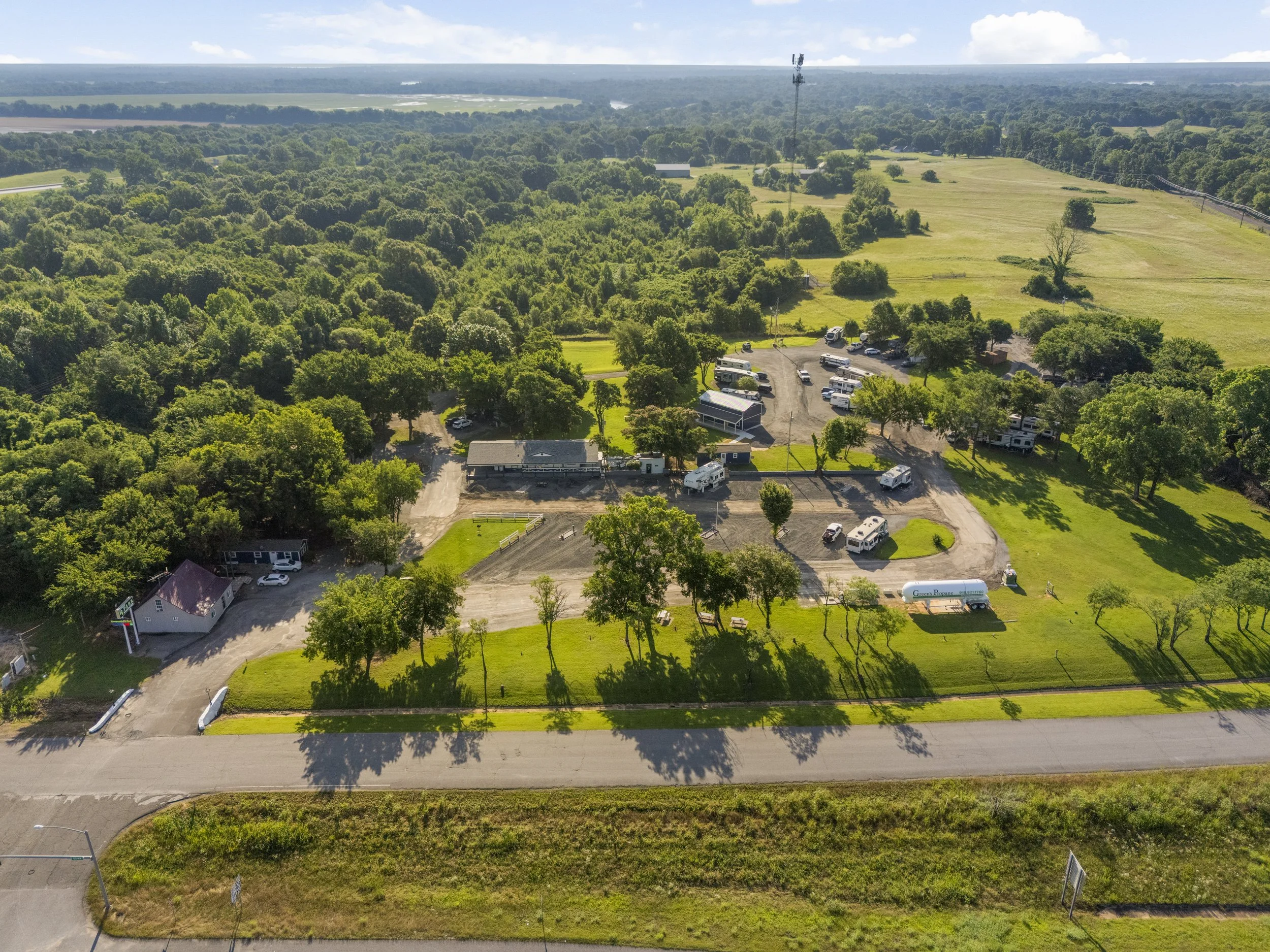 Aerial view of a rural area with dense trees, open grassy fields, and a small parking lot with vehicles and trailers. A road runs along the bottom of the image, and a large communication tower is visible in the distance.