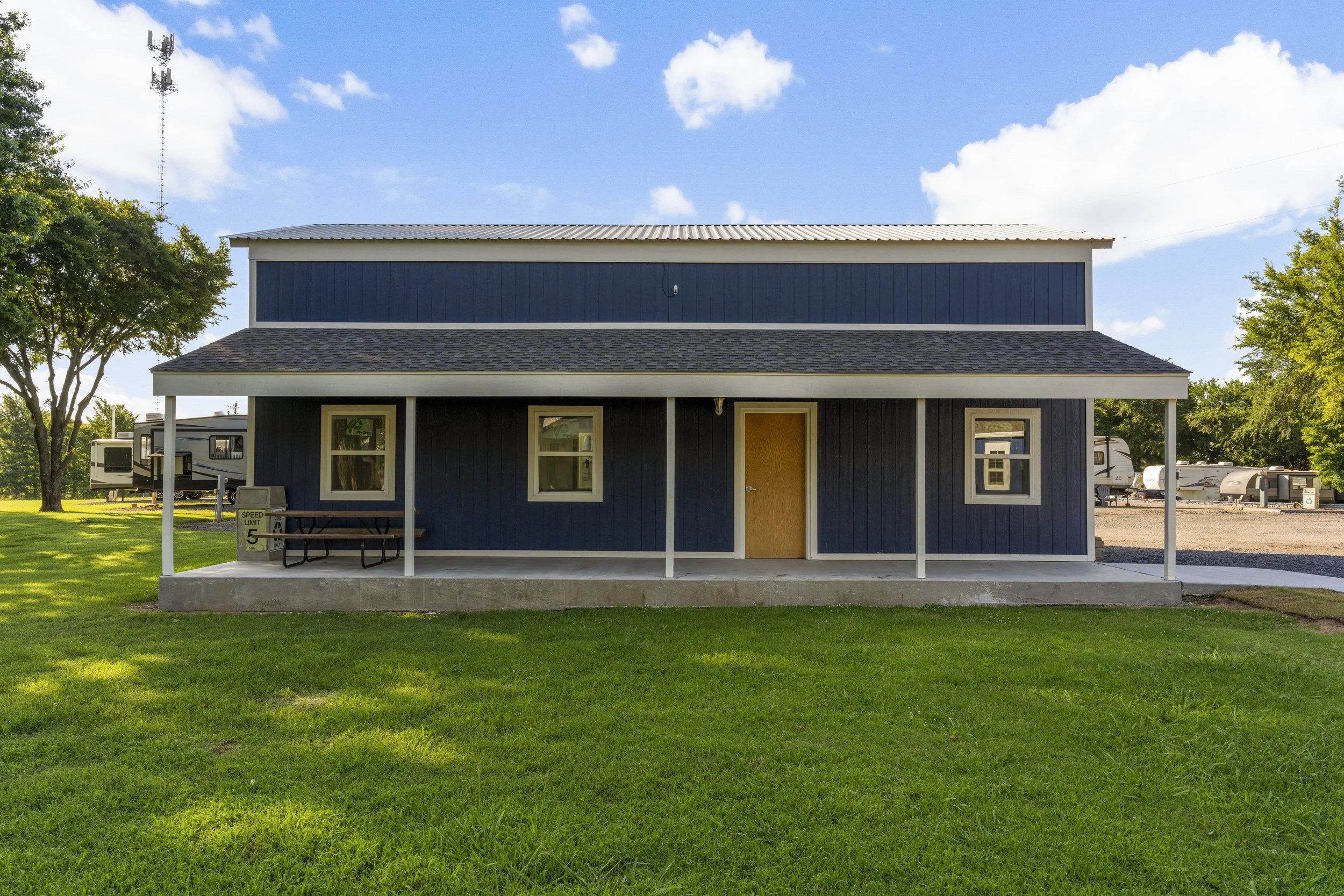 A two-story blue house with a porch, two white-framed windows, a wooden front door, surrounded by green grass, trees, and RVs in the background under a partly cloudy sky.