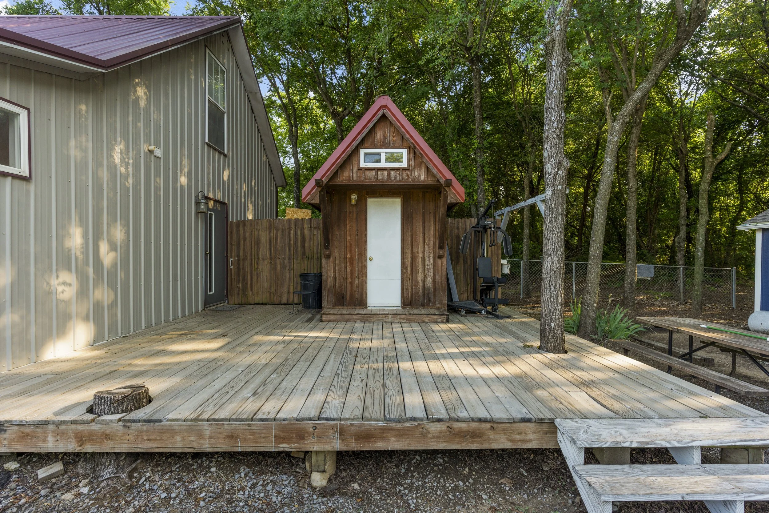 A backyard with a wooden deck and a small wooden shed with a red roof, surrounded by trees and a metal-sided building on the left.