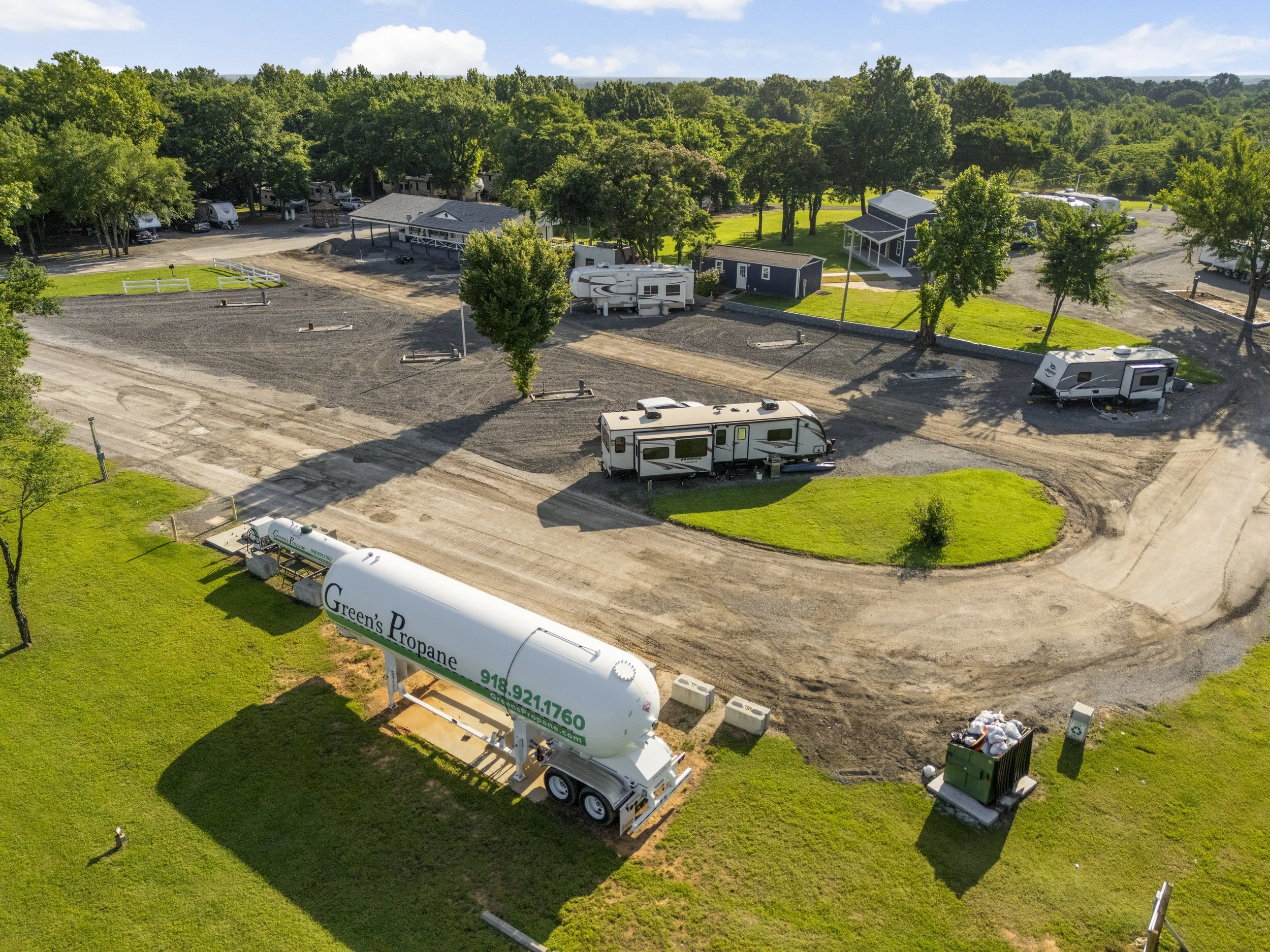 An aerial view of a rural property with trees, green lawns, a gravel parking area, and several parked trailers, including a large propane tank and some trash bins.