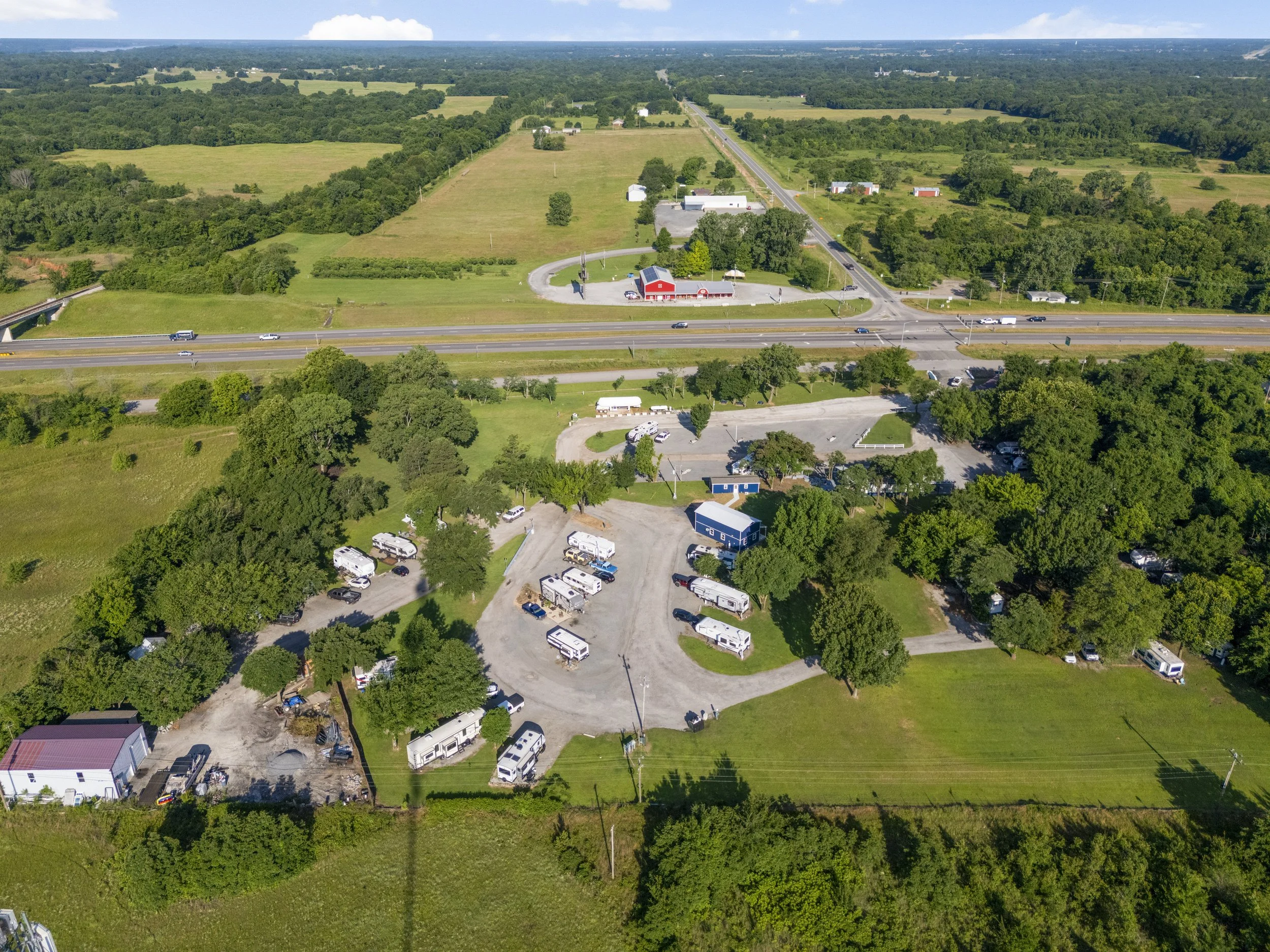 An aerial view of a rural area with green fields, trees, a highway with vehicles, and a small area with RVs and buildings.