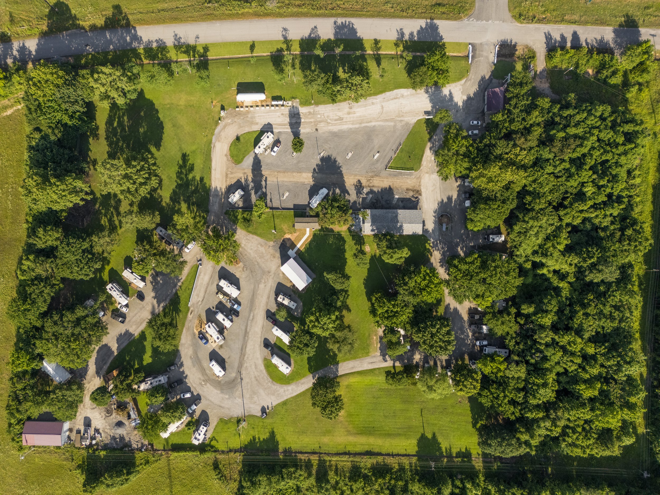 An aerial view of a rural area featuring a gravel driveway, several parked vehicles, green trees, a grassy area, and a few small structures, with a paved road at the top.