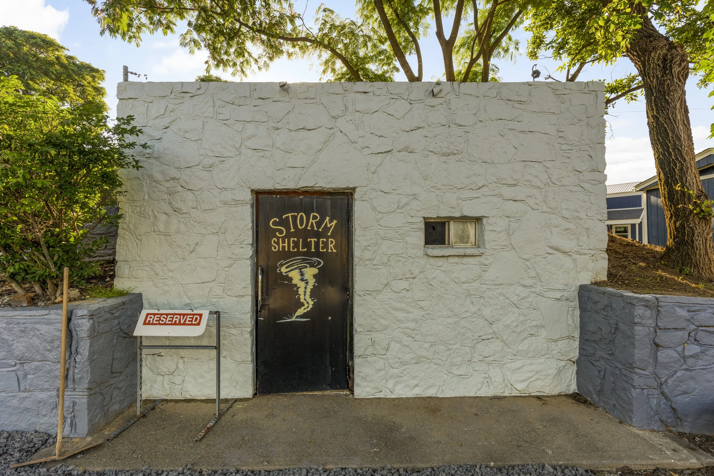 A small white stone building labeled 'Storm Shelter' with a tornado icon painted on the door. There is a 'Reserved' sign and a small shrub to the left. Trees surround the building, and sunlight filters through the leaves.