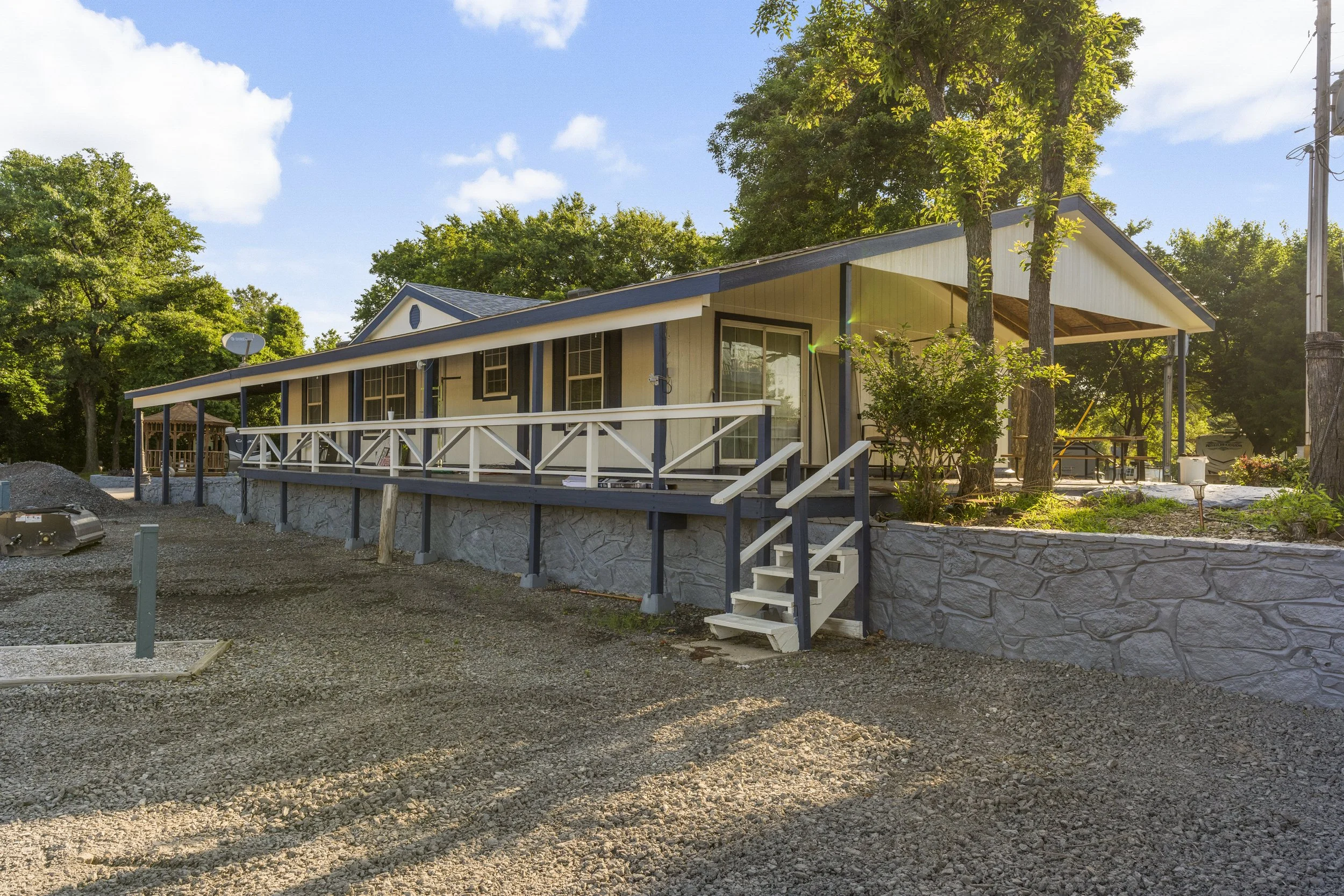 Newly constructed house with a sloped roof, elevated porch with white railing, and stairs, surrounded by trees and construction materials at a sunny outdoor site.