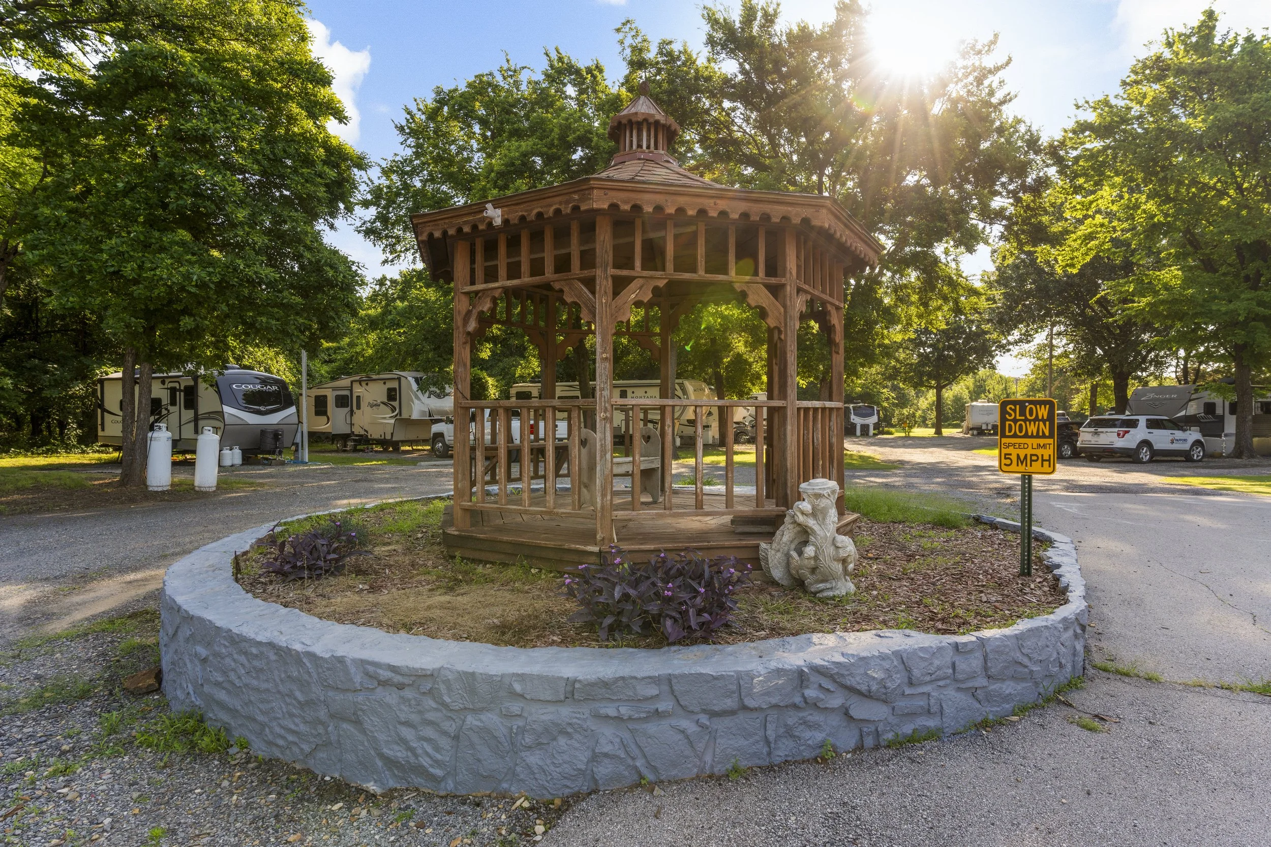 A wooden gazebo in a park with trees and a parking area behind it. The sun is shining through the trees, creating a lens flare. There is a sign that says 'Slow Down 5 MPH'.
