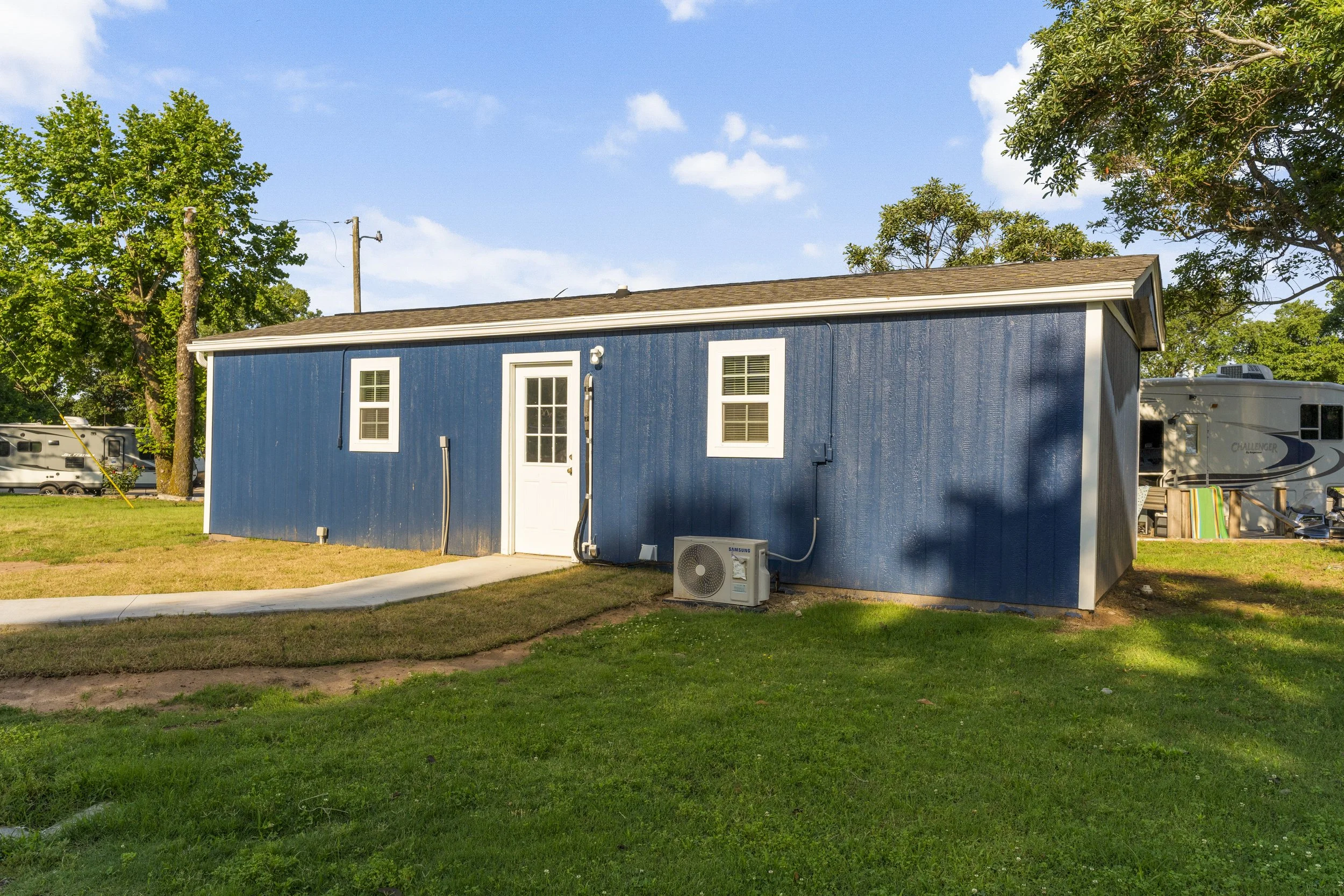 A small blue house with white trim, two windows, and a door, located on a grassy area with trees and a clear blue sky in the background.