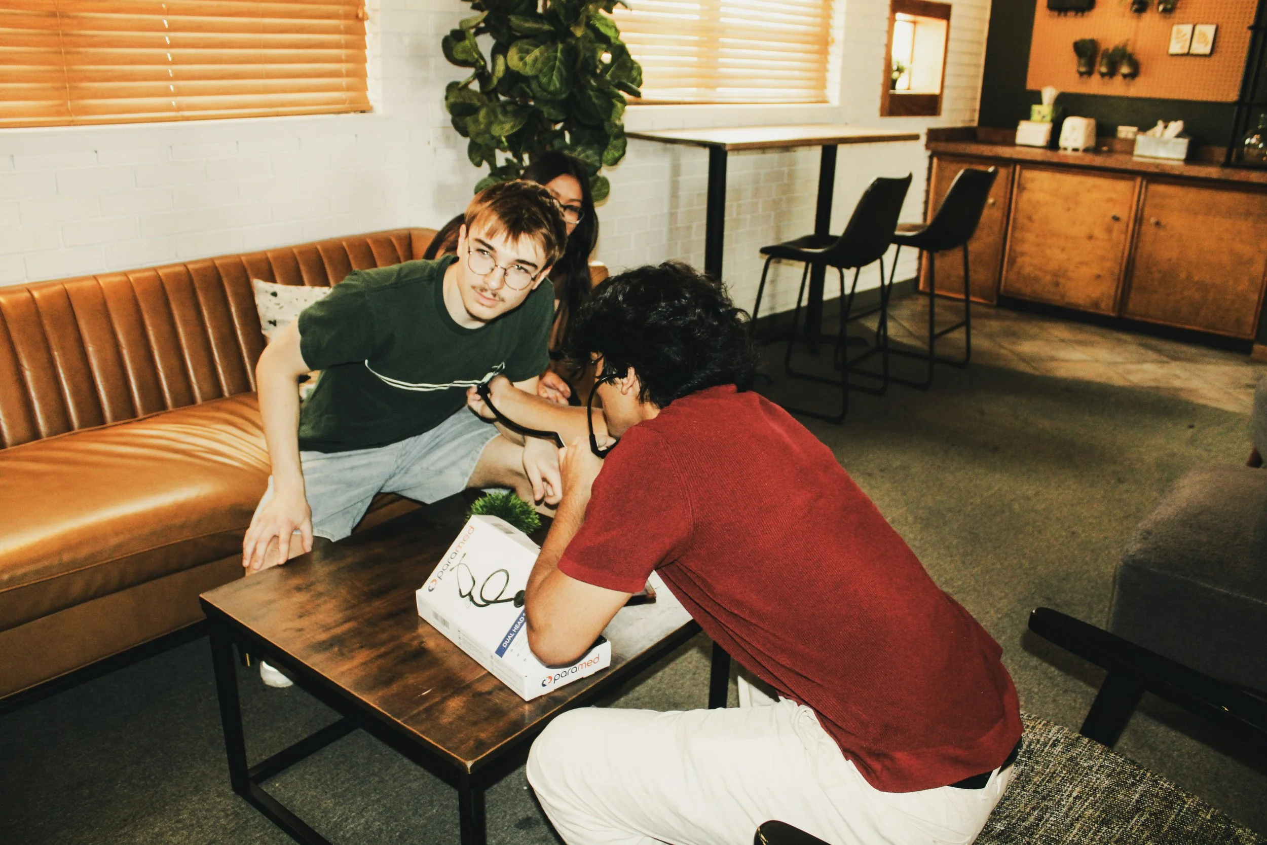 A young man with glasses and a green shirt leaning on a table, talking to an older woman with glasses and black hair, wearing a red shirt, in a cozy room with wooden cabinets, a sofa, and large windows.