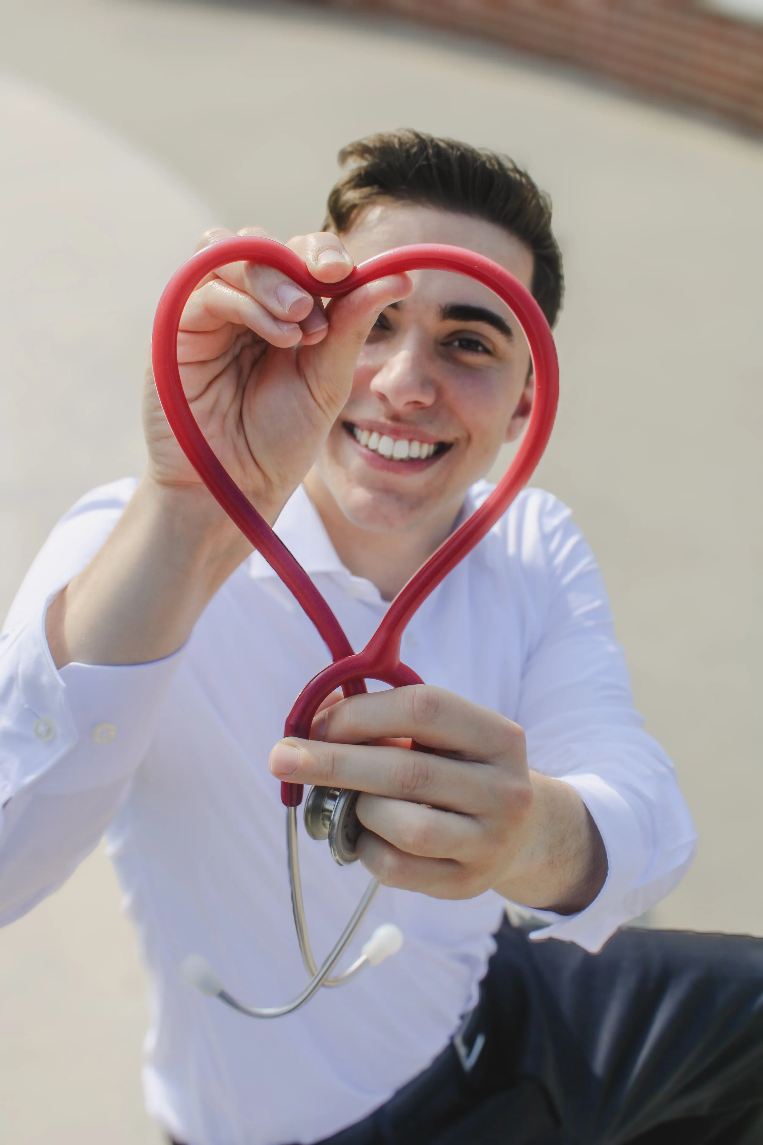 A young man in a white shirt smiling and holding a red stethoscope shaped like a heart, looking through the heart shape at the camera.