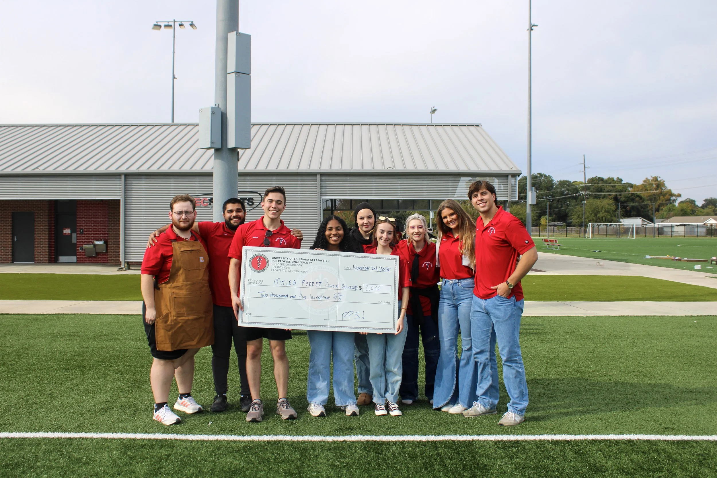 A group of nine young adults smiling and posing outdoors on a football field, holding a large check for $2,500 made out to Miles Perret Cancer Services, with a school building and football field in the background.