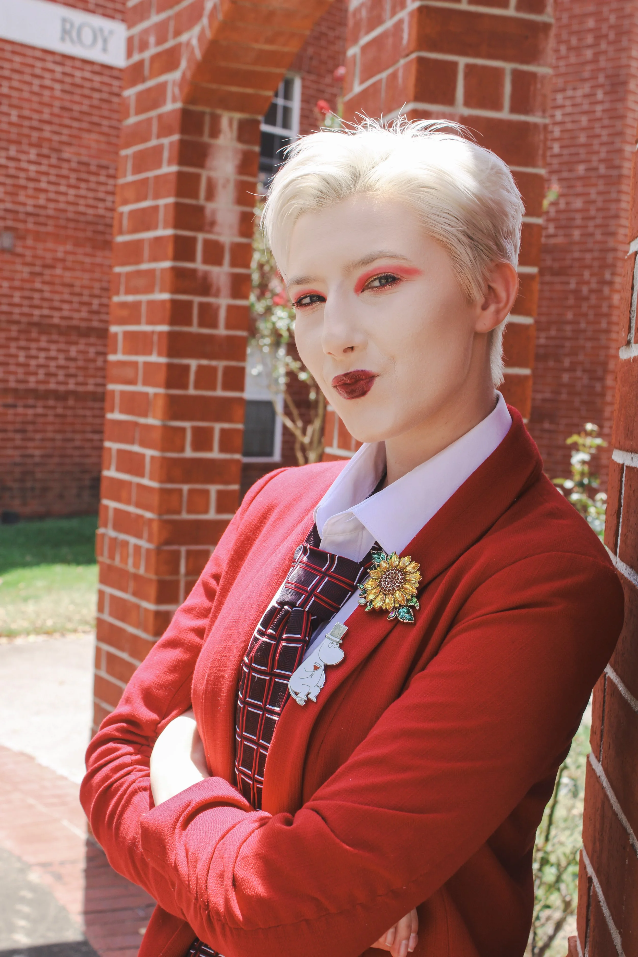 A woman with platinum blonde hair with darker roots, wearing dark lipstick, a black leather jacket, earrings, and a necklace, standing outdoors in a forested area.