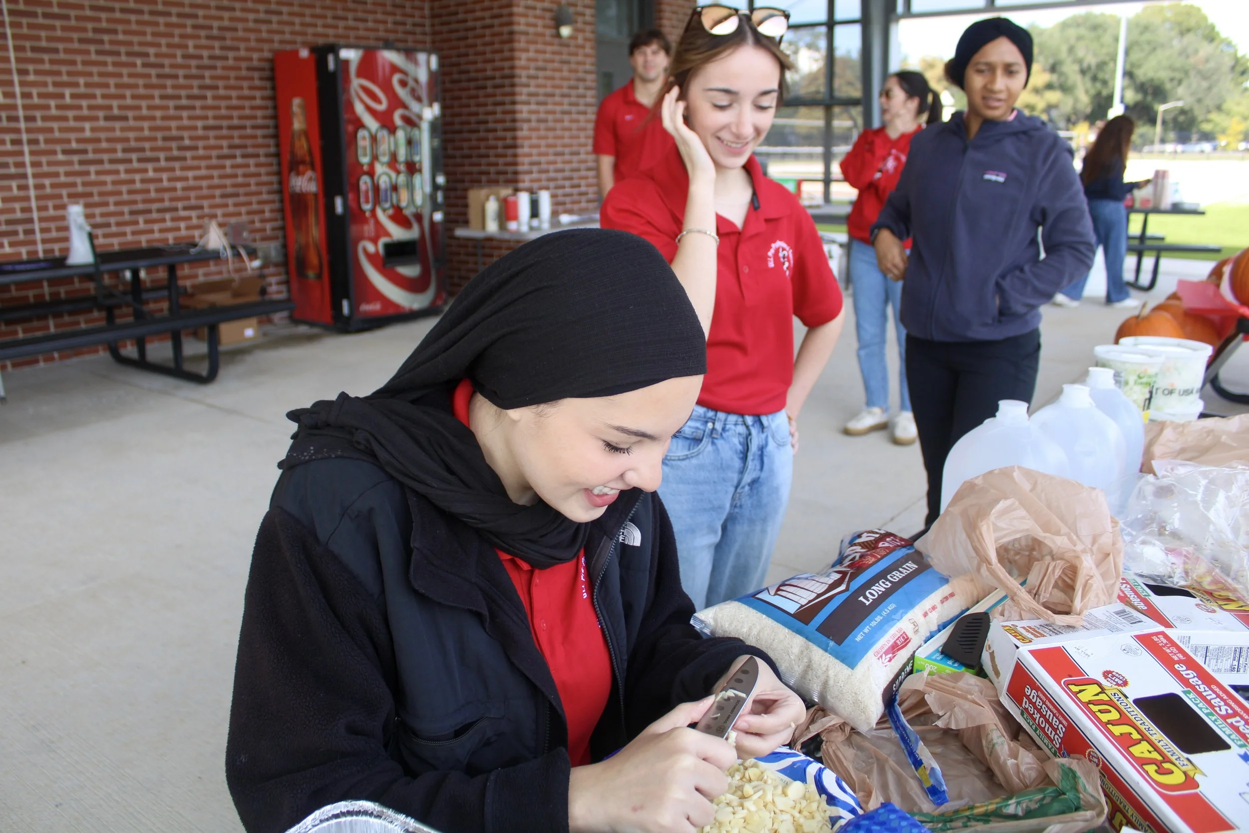 Group of young people at a food drive, organizing supplies and food items at a table outside a brick building with a large glass window.