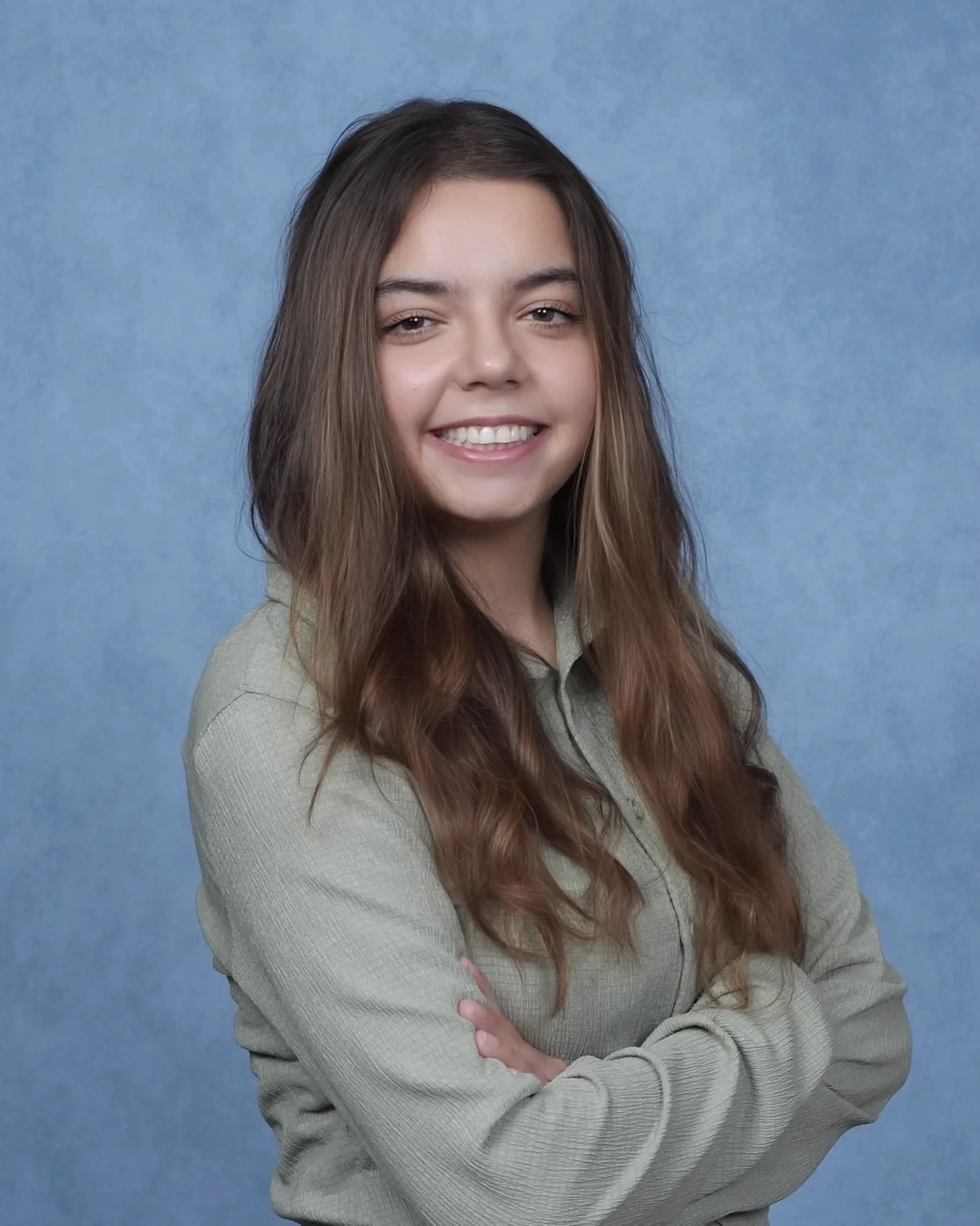 A young woman with long brown hair and a big smile, standing with arms crossed against a blue background, wearing a beige sweater.