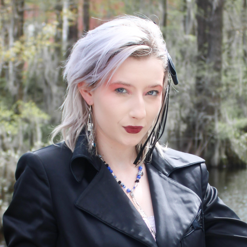 A woman with platinum blonde hair with darker roots, wearing dark lipstick, a black leather jacket, earrings, and a necklace, standing outdoors in a forested area.