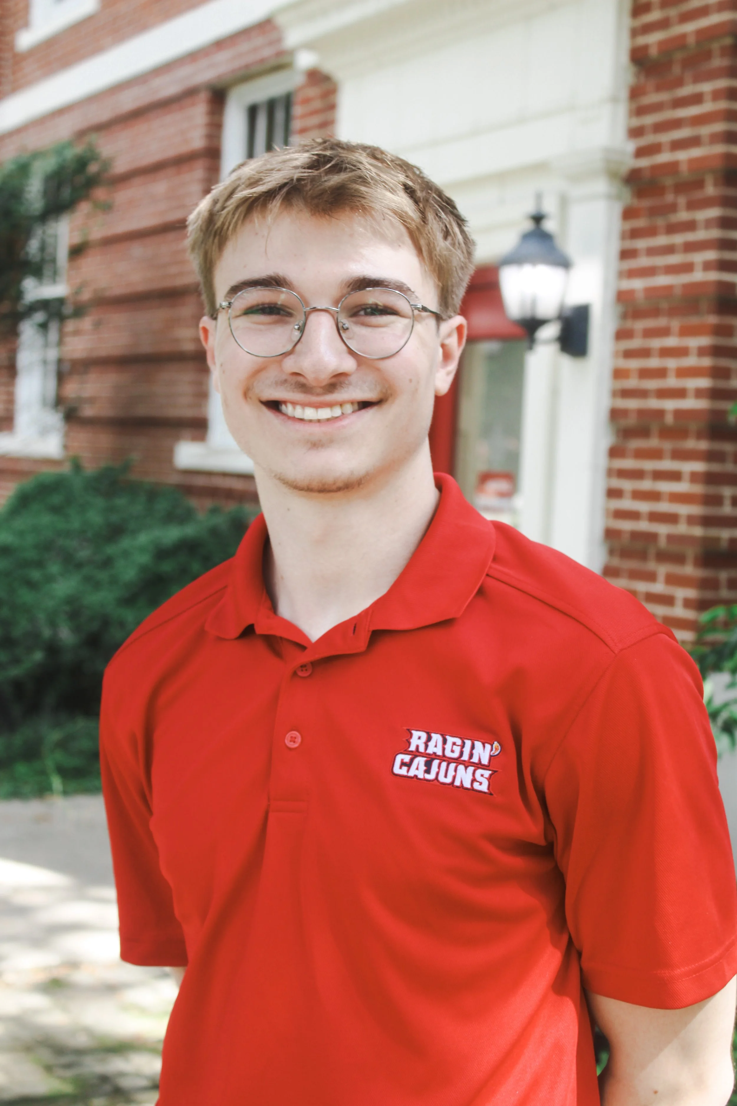Headshot of a young man with light brown hair, glasses, and a smile, wearing a light checkered dress shirt.
