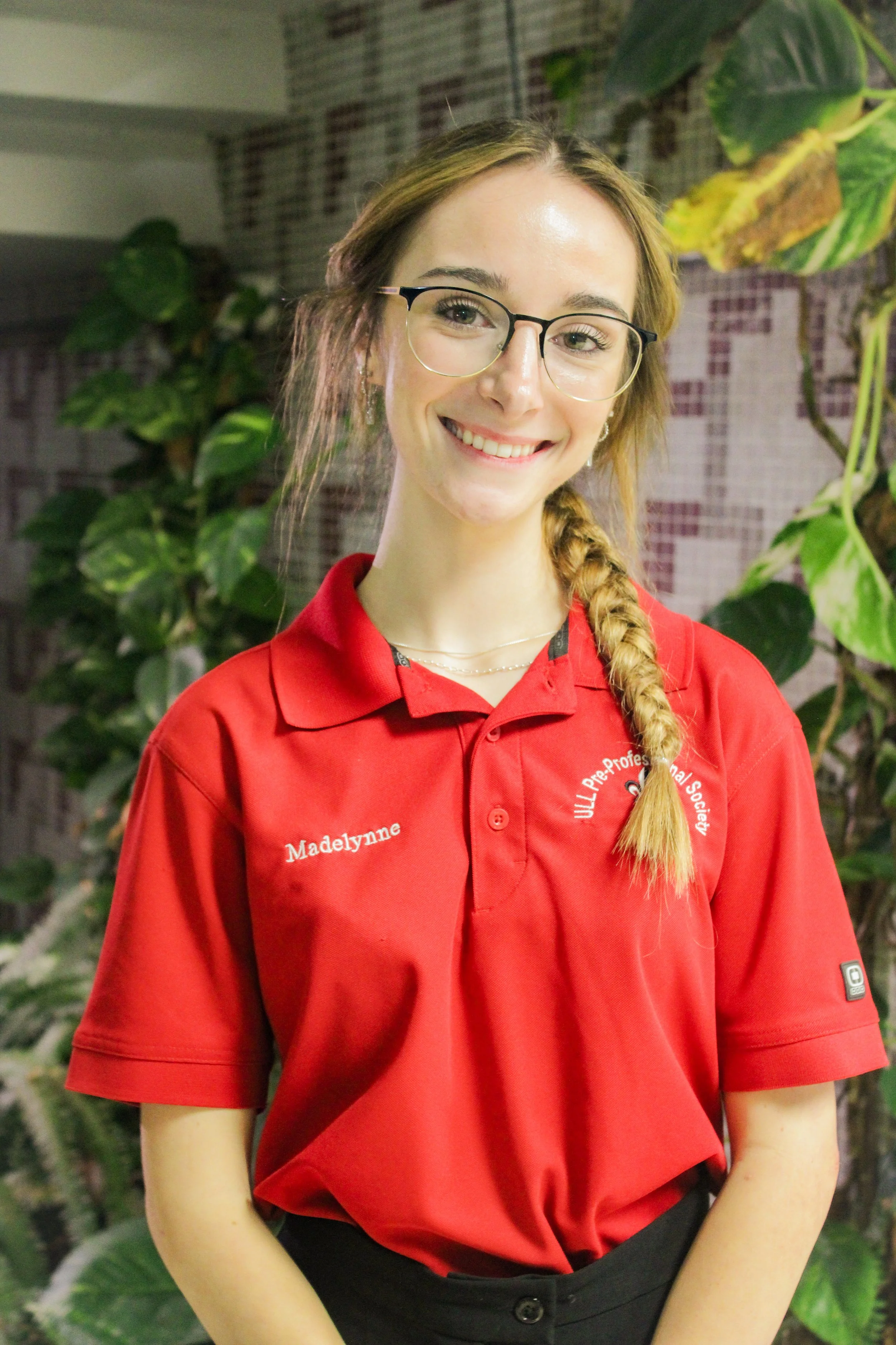A woman with glasses and a side braid, wearing a red shirt with embroidered text, smiling at the camera, surrounded by green plants.