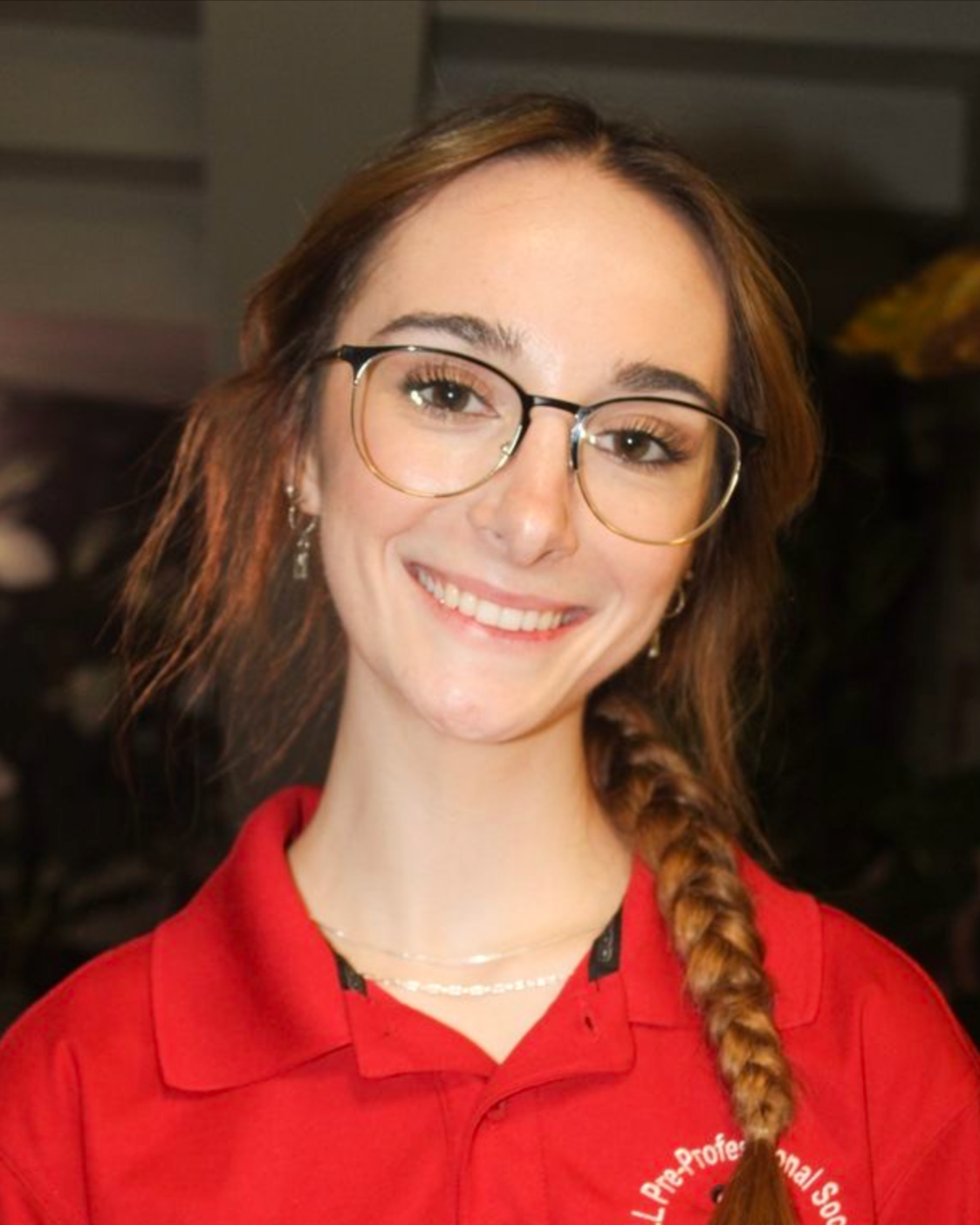 A woman with glasses and a side braid, wearing a red shirt with embroidered text, smiling at the camera, surrounded by green plants.